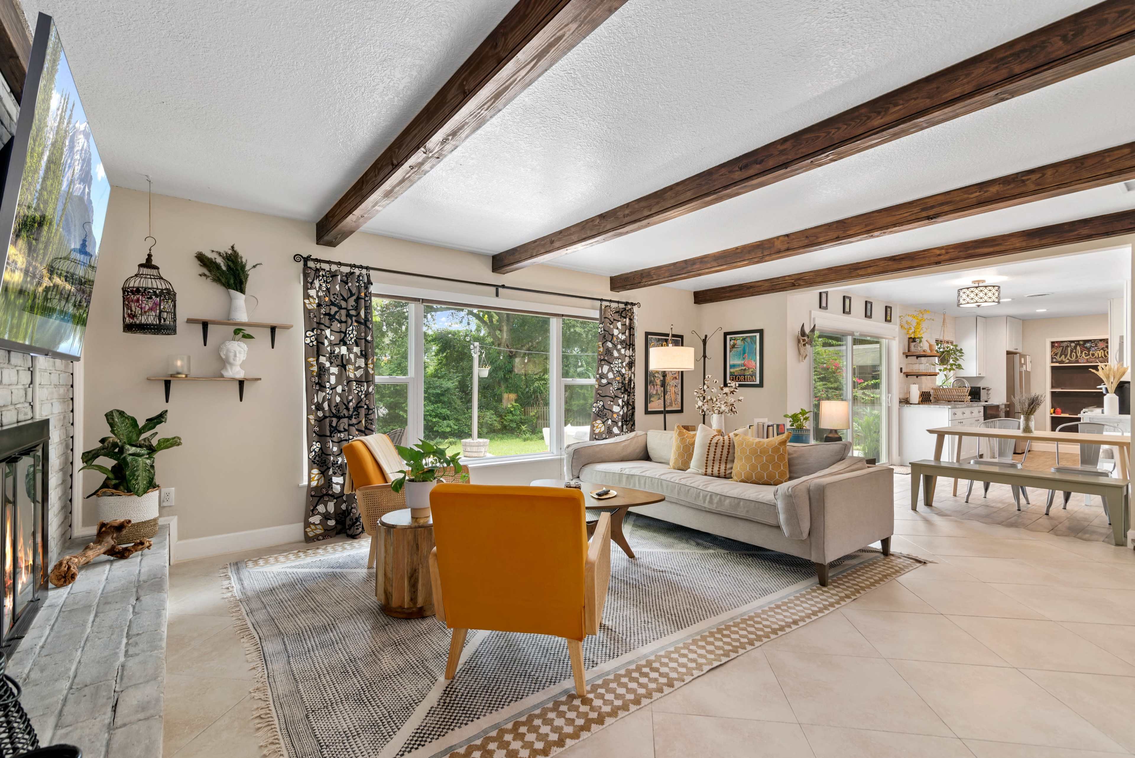 A modern living room features a gray sofa, yellow accent chairs, and large windows overlooking a garden, with wooden beams on the ceiling.