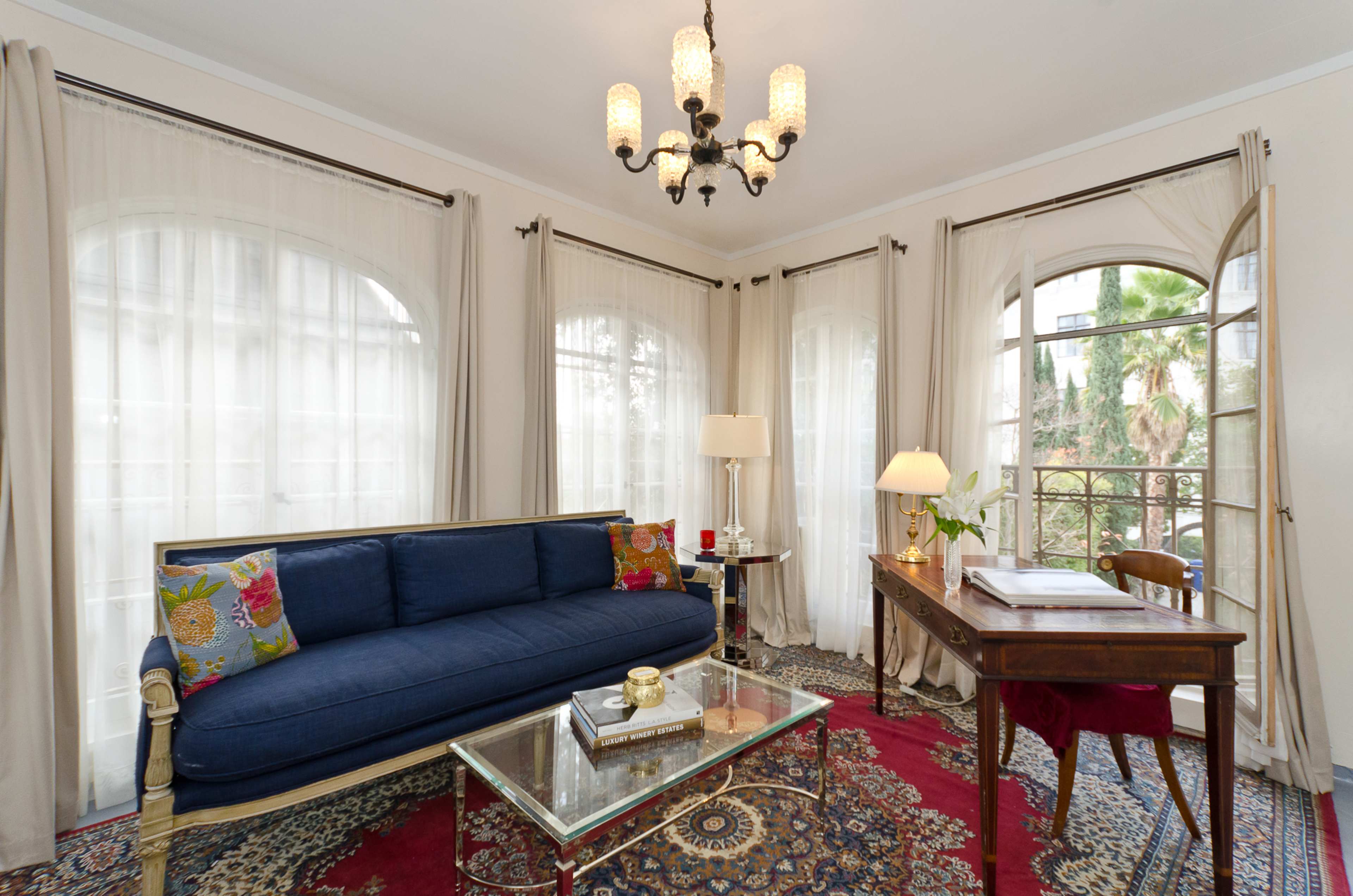 The room features a blue sofa, a glass coffee table, and a wooden desk beside large windows with sheer curtains, offering a view of greenery outside.