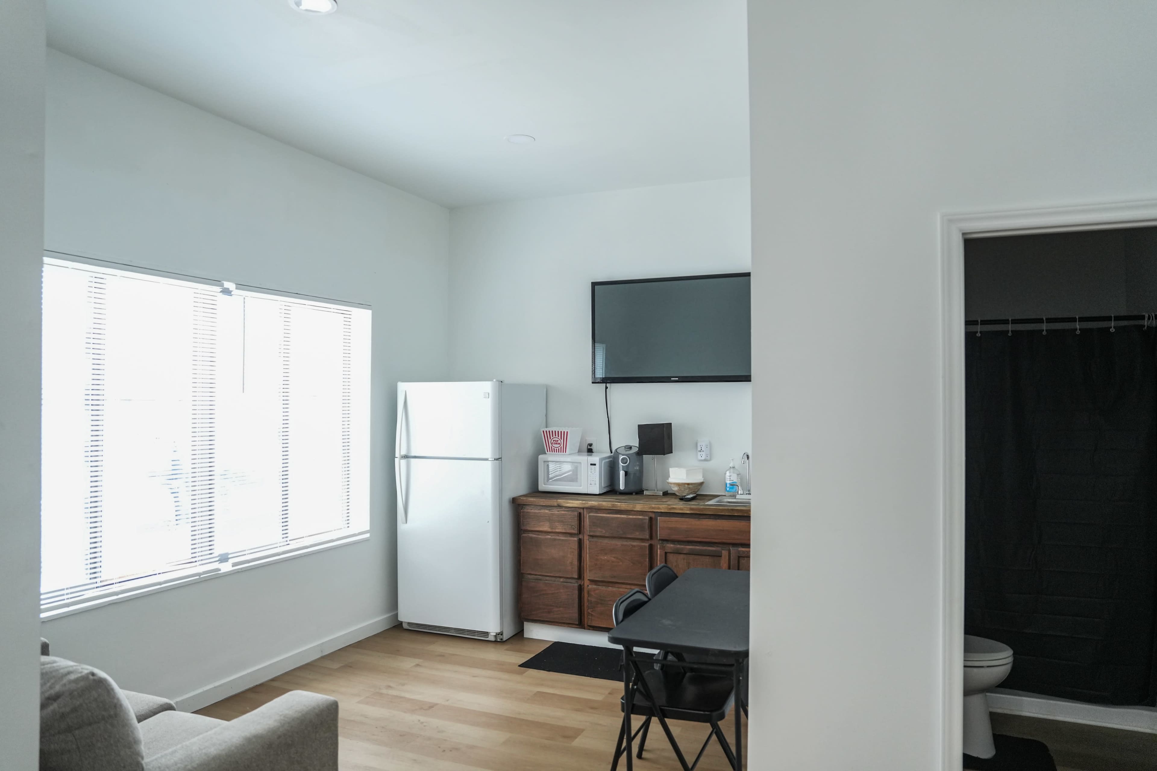 A small kitchen area features a white refrigerator, a wall-mounted television, and a dining table in a minimalistic living space with a nearby bathroom.