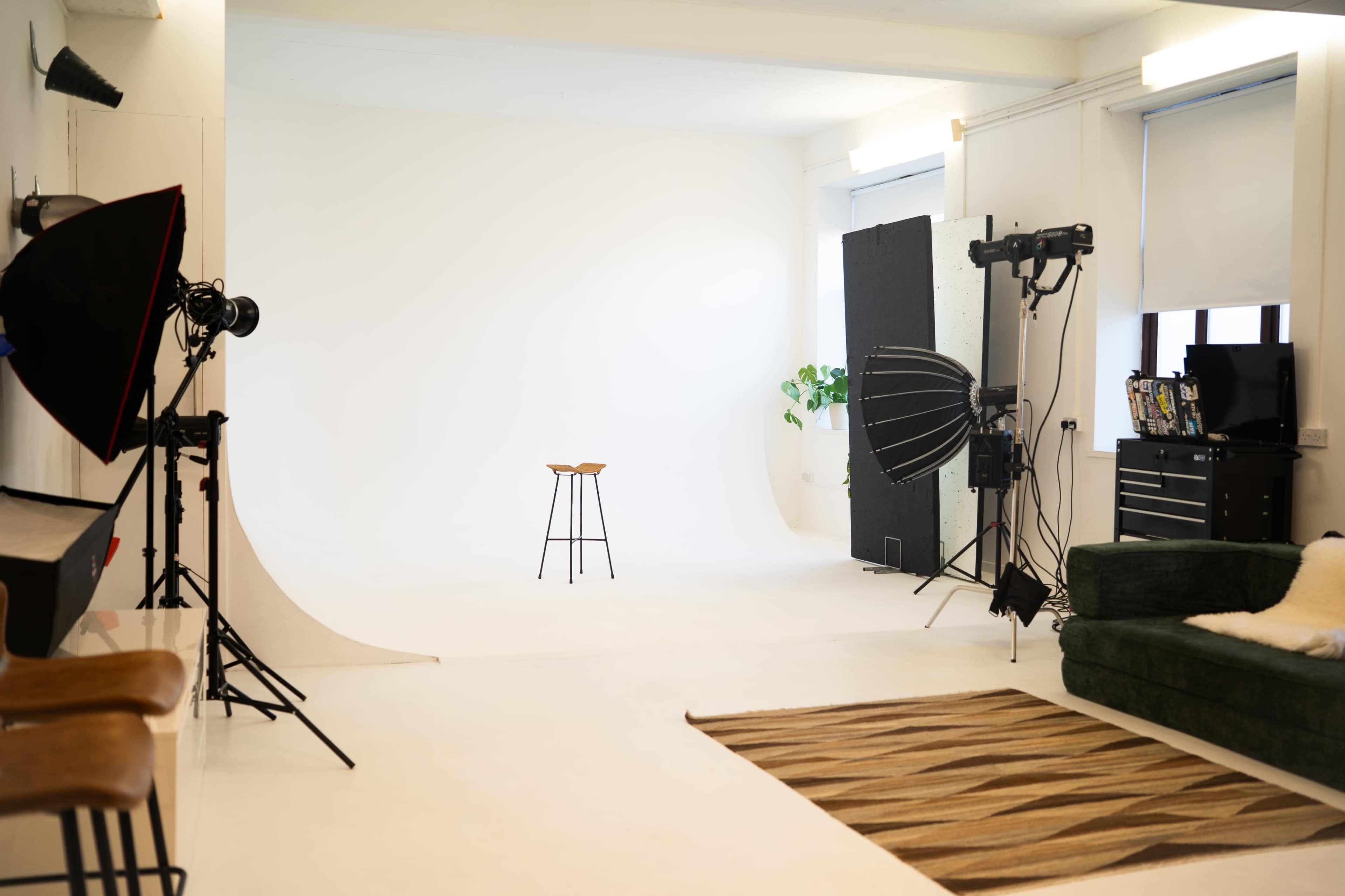 The image shows a minimalist photography studio with a single wooden stool on a white backdrop, surrounded by various photography equipment and a green couch.