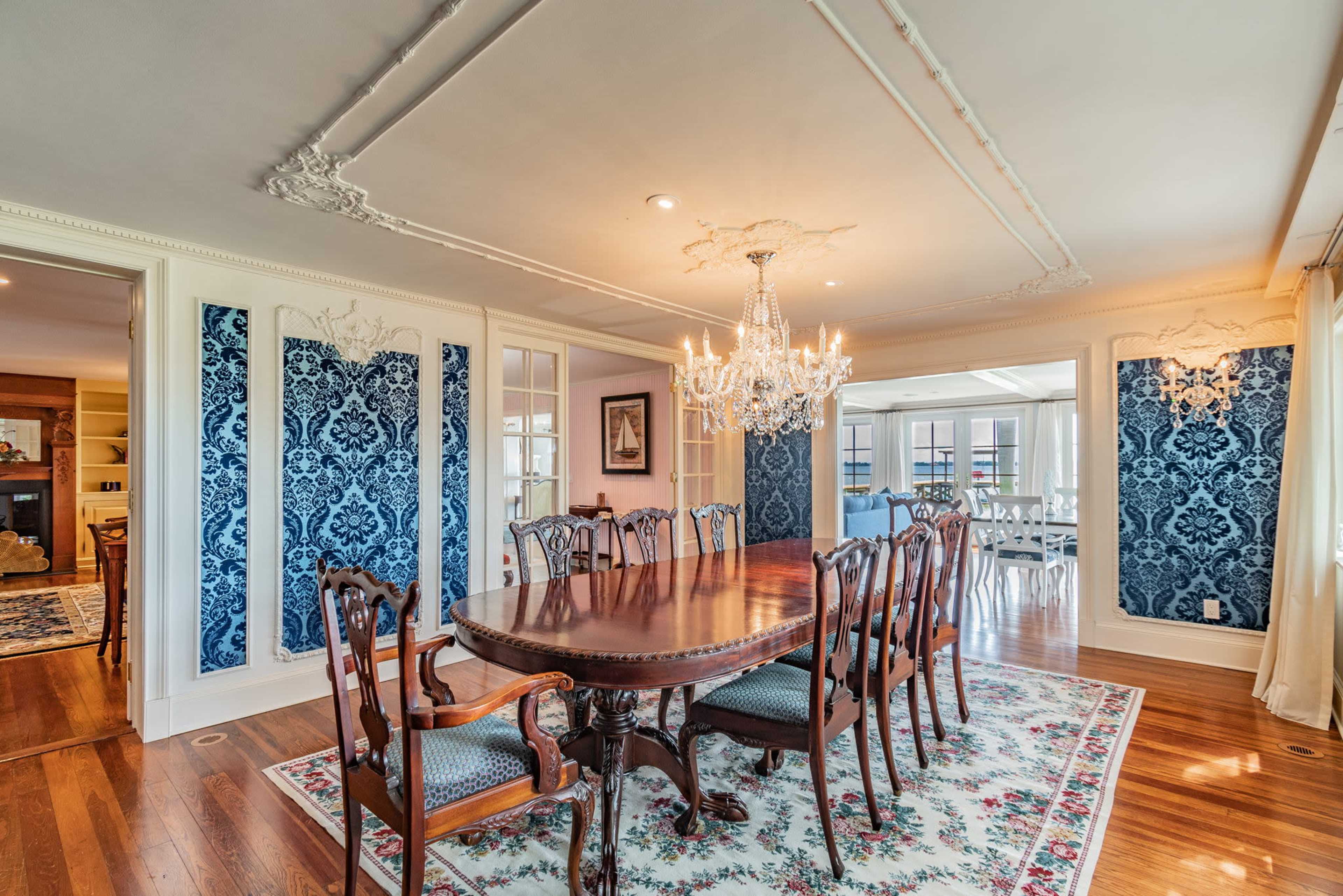 The dining room features a large wooden table surrounded by twelve chairs, illuminated by a chandelier and accented by blue patterned wallpaper and floral carpeting.