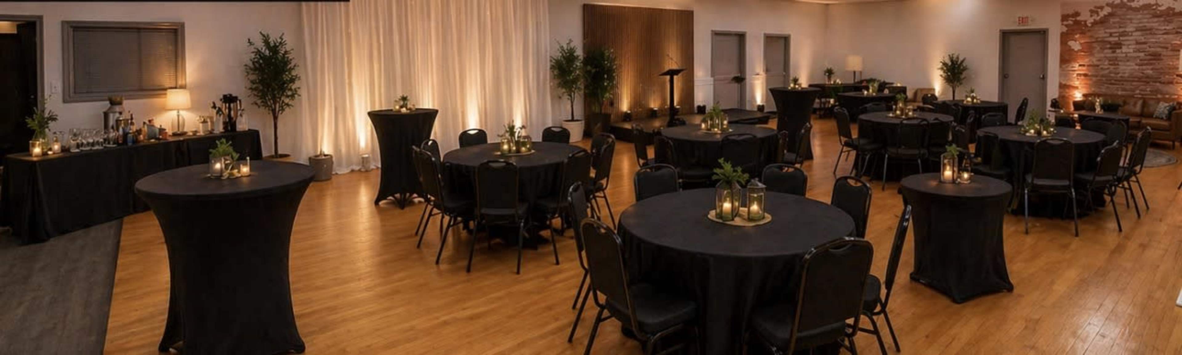 The image shows a spacious event venue with round black tables, each adorned with small centerpieces, and ambient lighting creating a warm atmosphere.