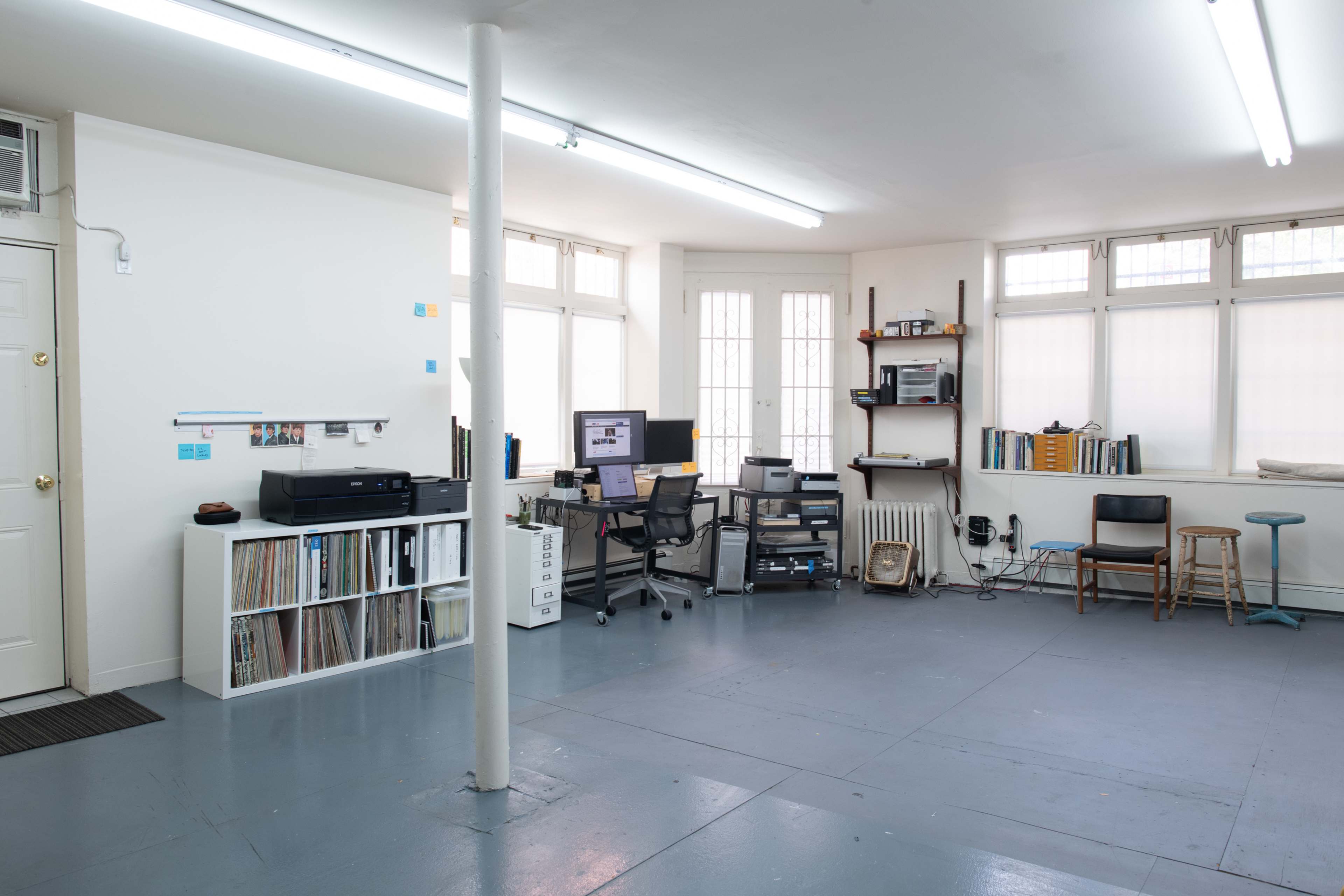 The image shows a spacious, minimally furnished office or studio with a desk, computer setup, shelves, and a printer against the walls, featuring large windows that provide natural light.