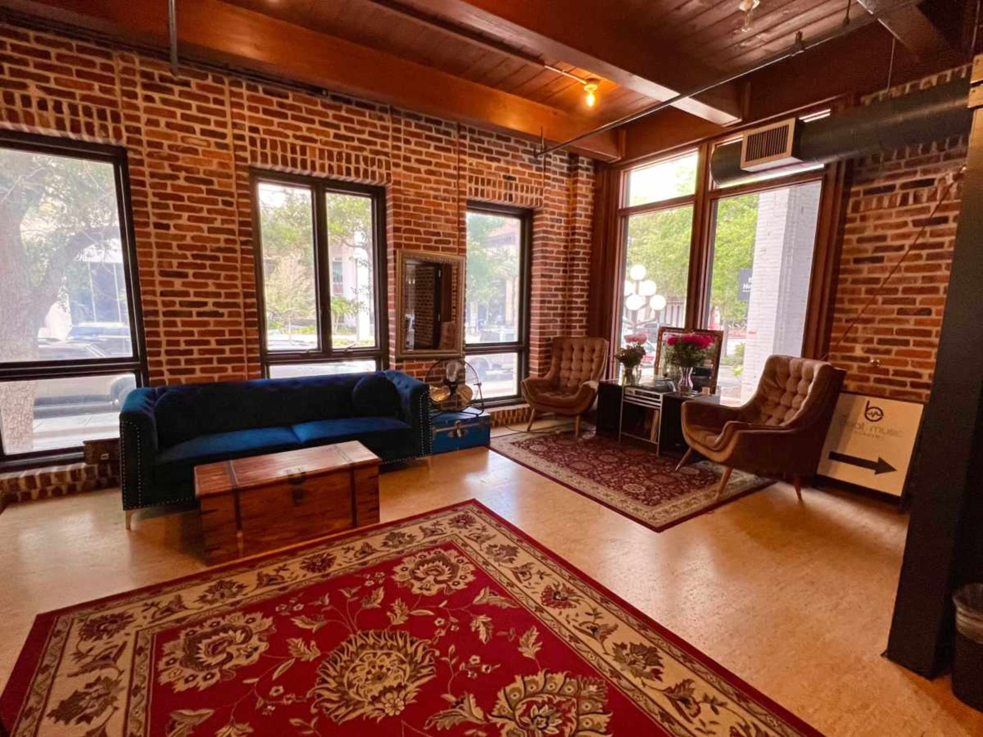 The image shows a cozy lounge area with exposed brick walls, two brown chairs, a blue couch, and a coffee table on a patterned rug.