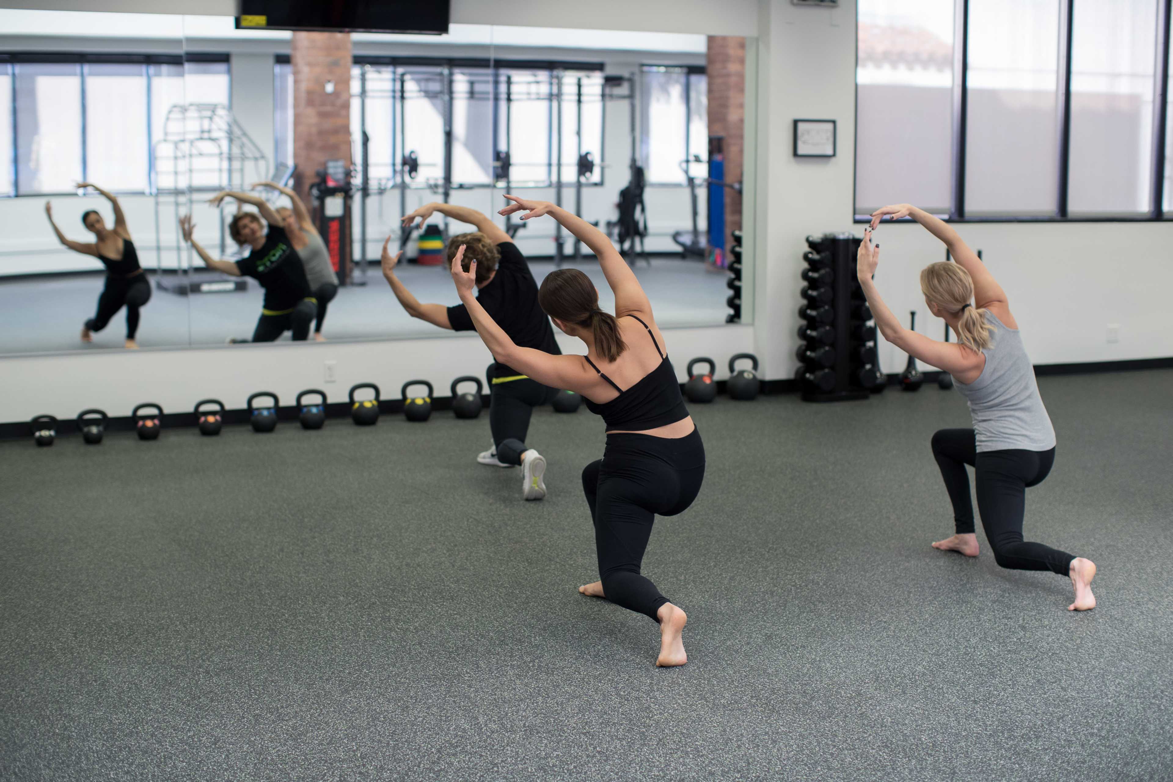 Three women perform a dance routine in a mirrored fitness studio with exercise equipment in the background.