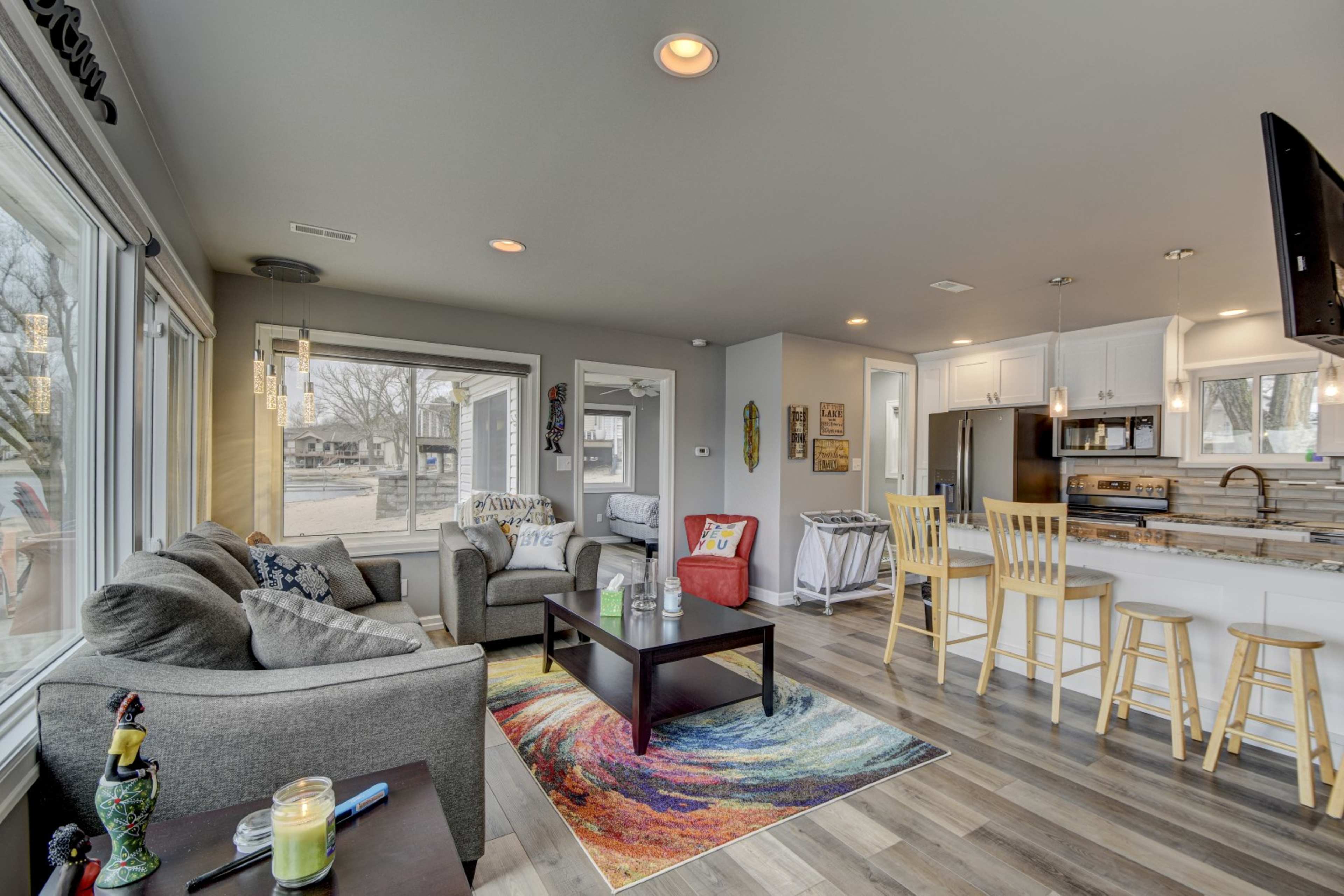 The living room features a gray couch, a colorful rug, and a kitchen bar with four stools, with large windows offering a view of the outdoors.
