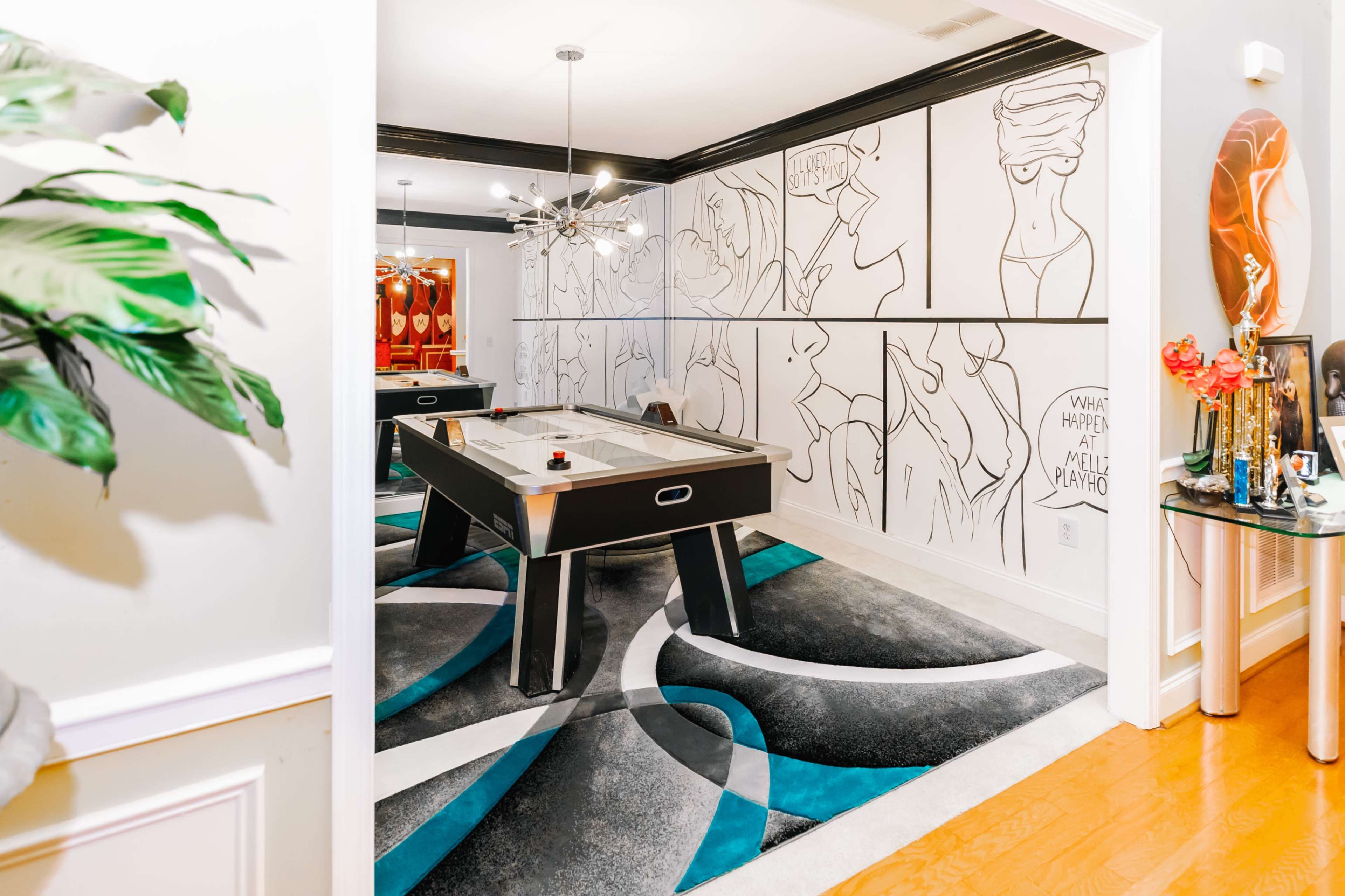 The image shows a modern game room featuring an air hockey table, a vibrant area rug, and large wall art with bold line drawings.