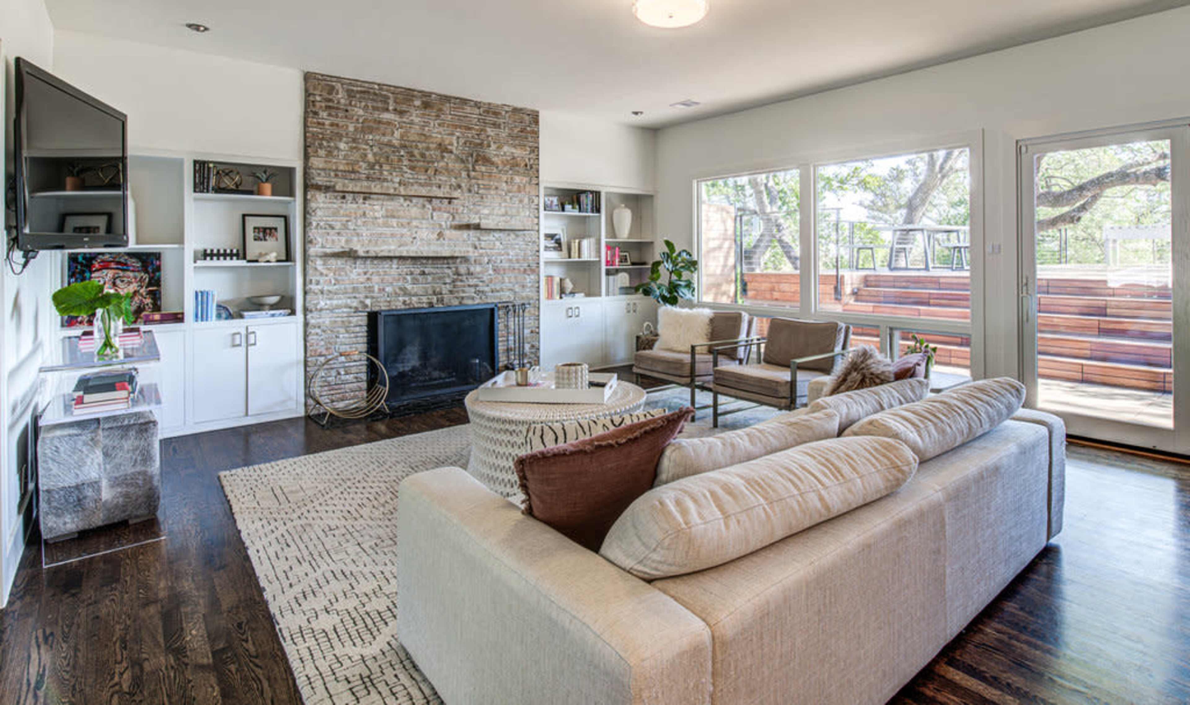 The image shows a modern living room with a large beige sofa, a circular coffee table, a stone fireplace, built-in shelves, and large windows letting in natural light.