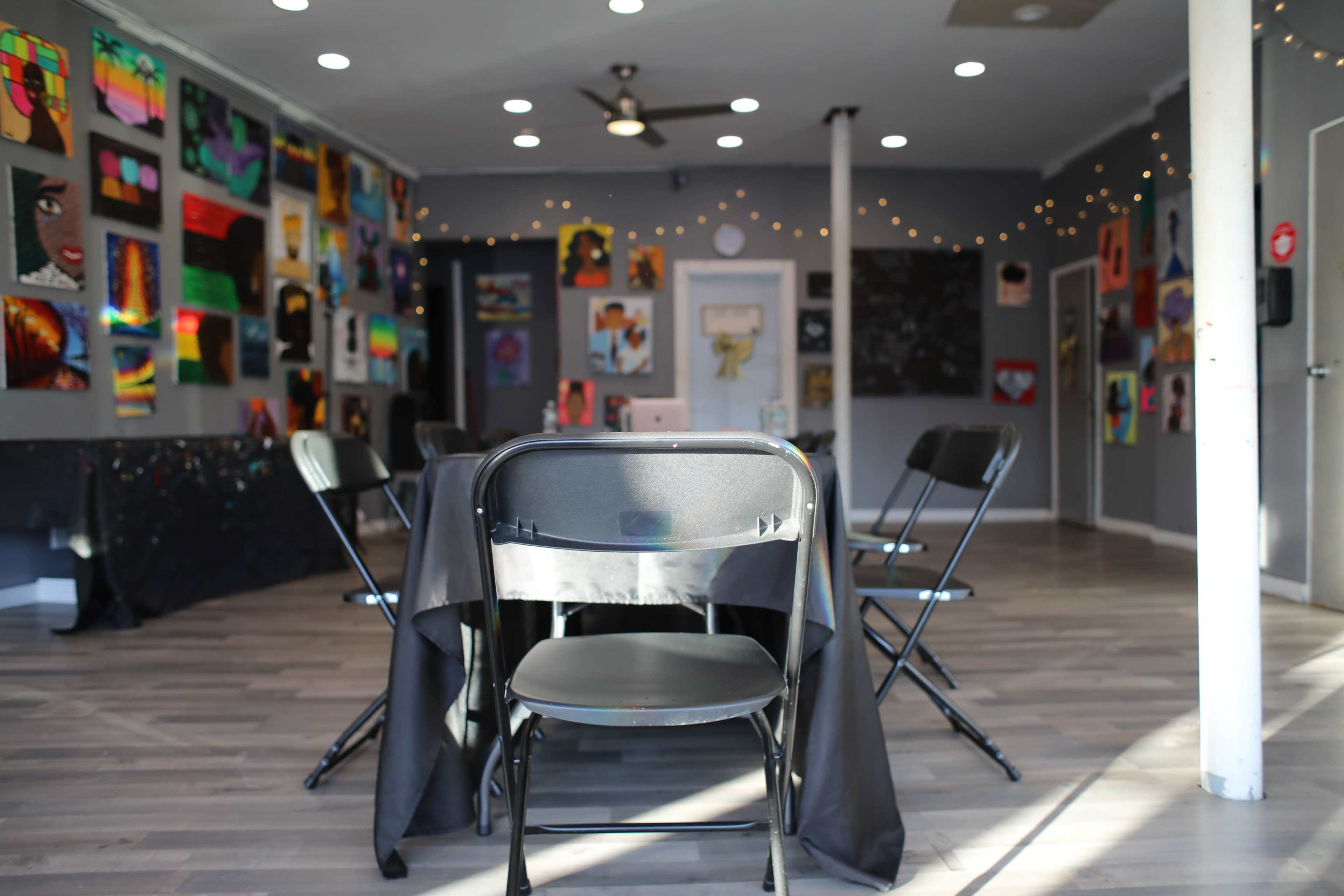 A spacious room with folding chairs arranged around a table, featuring vibrant artwork displayed on the walls.