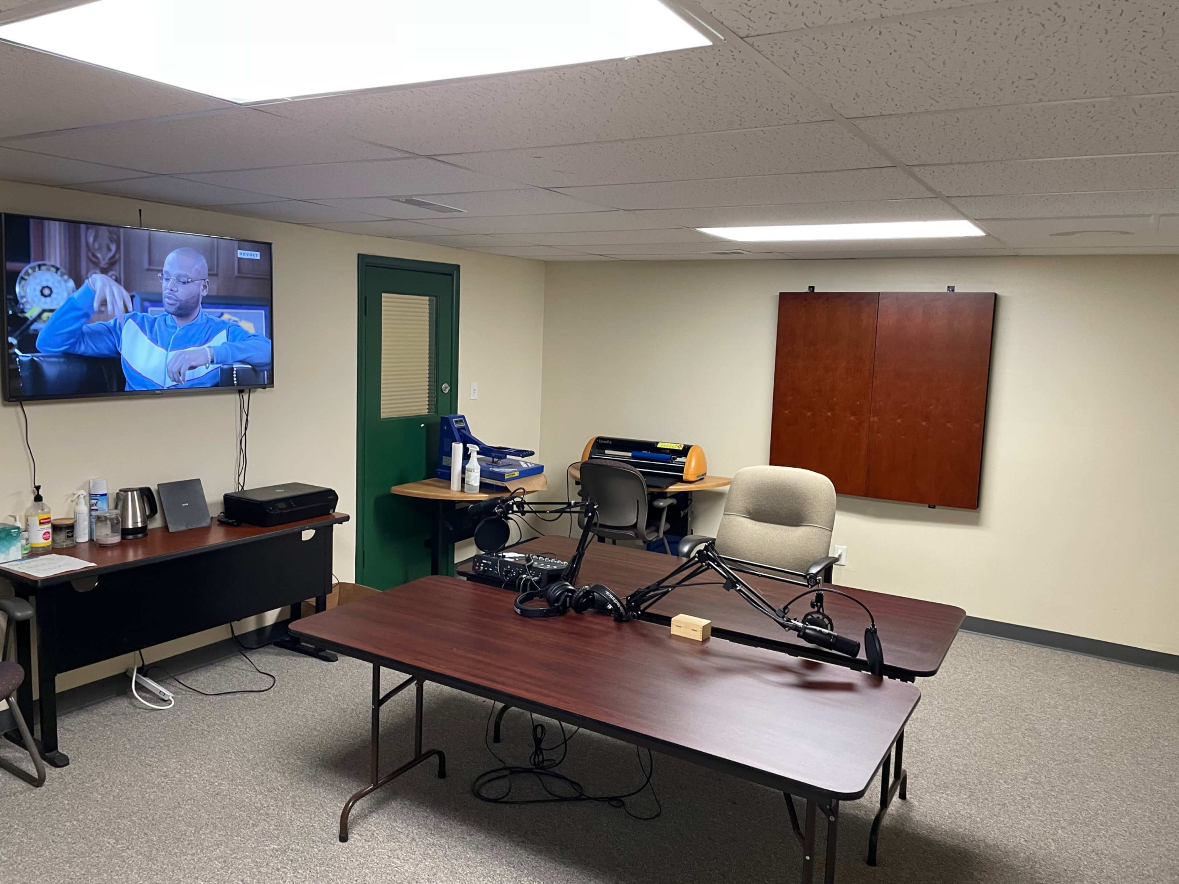 The image shows a sparsely furnished meeting room with a large table, a microphone setup, and a television displaying a scene.