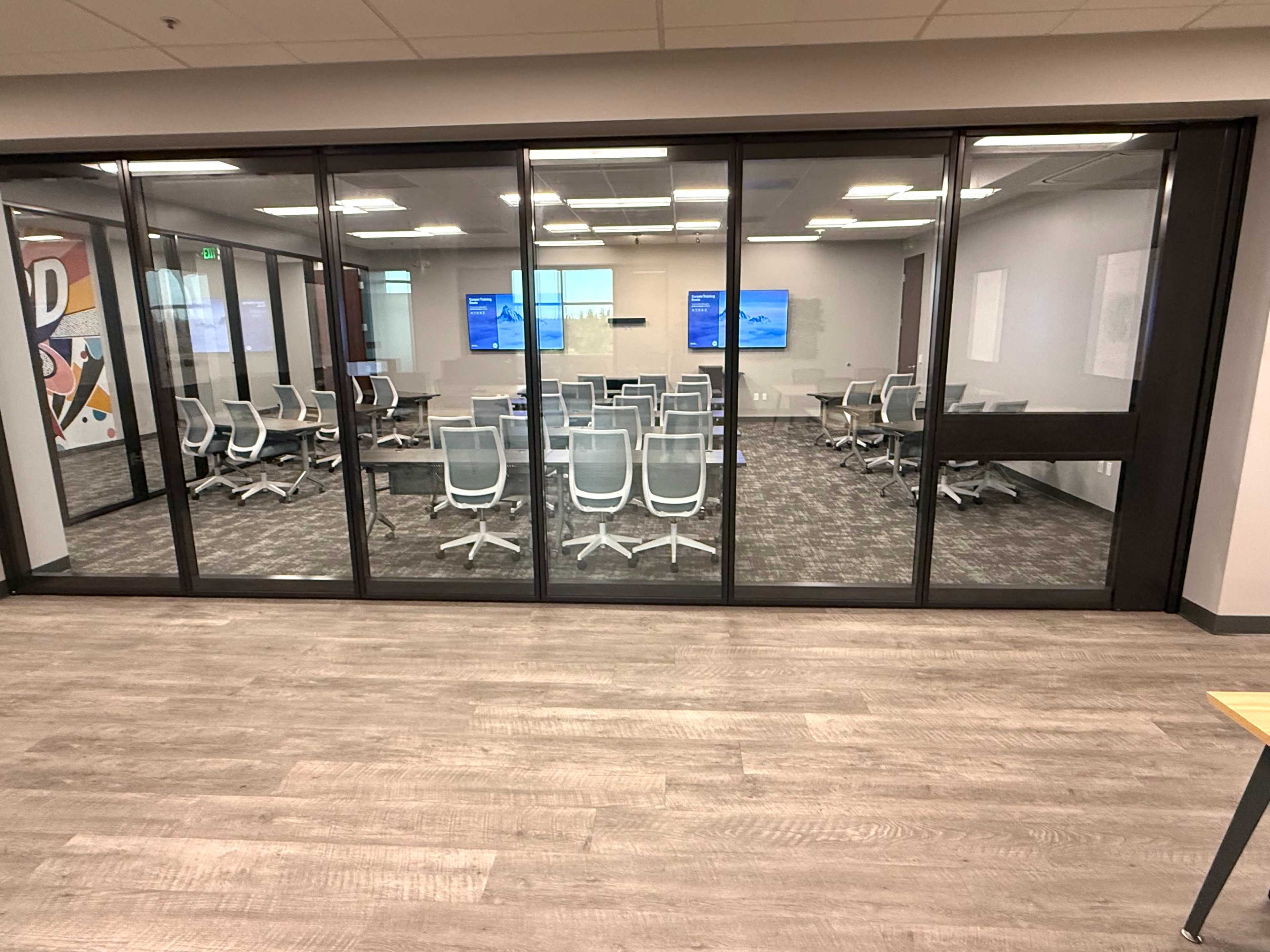 The image shows a modern conference room with glass walls, featuring white chairs arranged around a central table and two large screens visible on the far side.