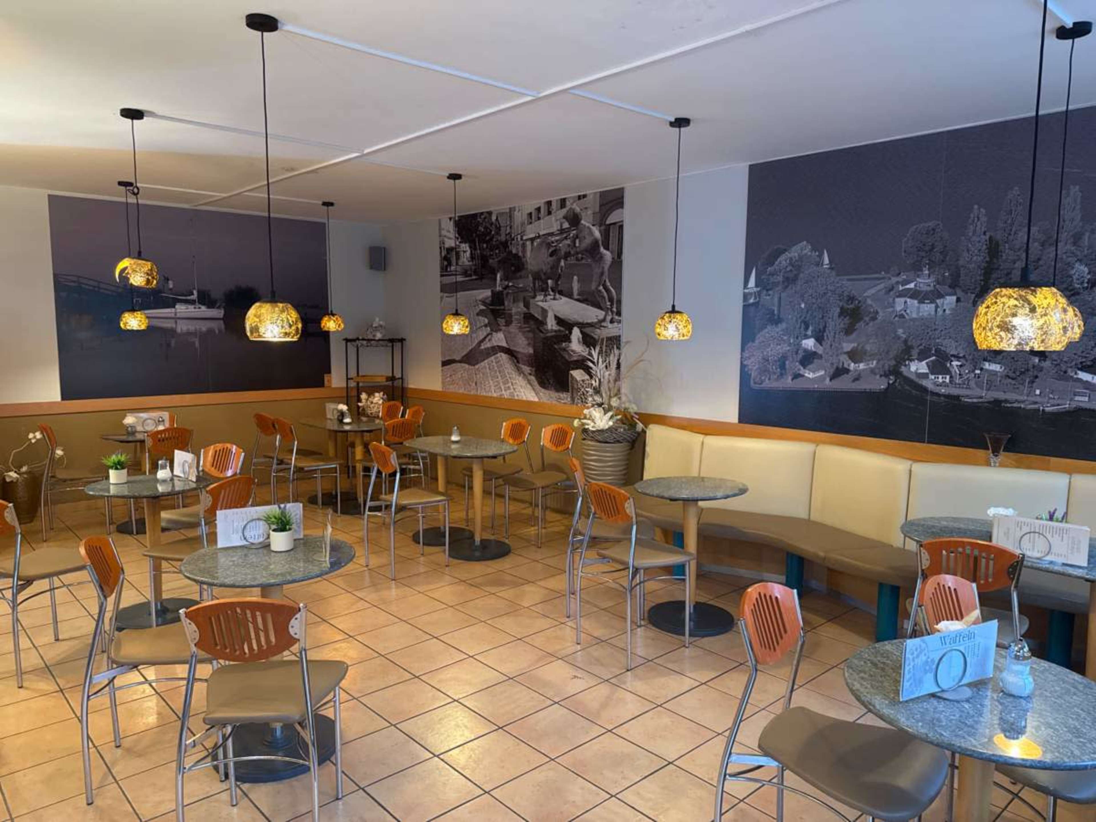 The image shows a dining area of a café with round tables, orange chairs, and large wall murals, illuminated by hanging pendant lights.