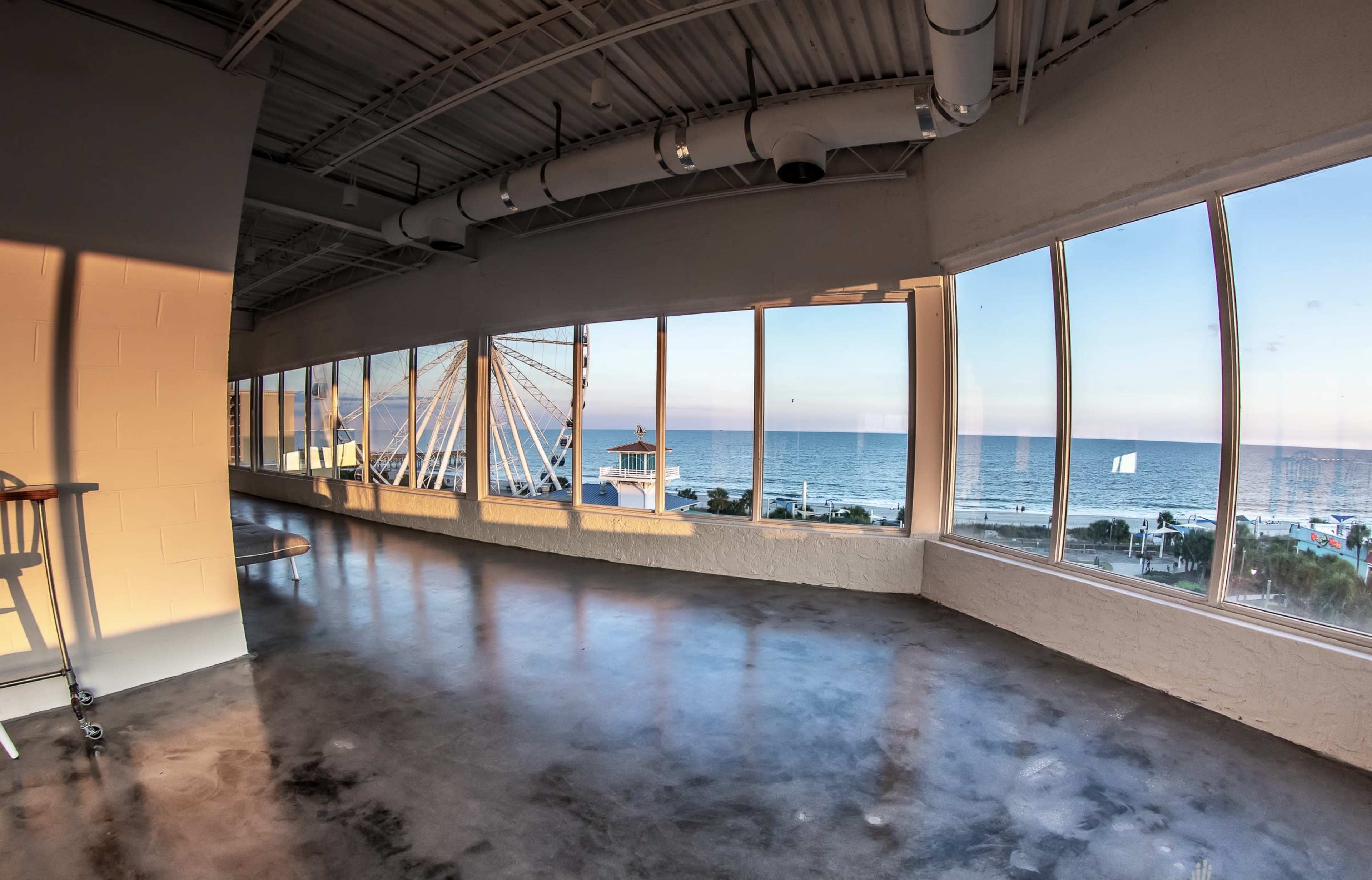A modern, empty room with large windows overlooking a beach and a Ferris wheel in the distance.