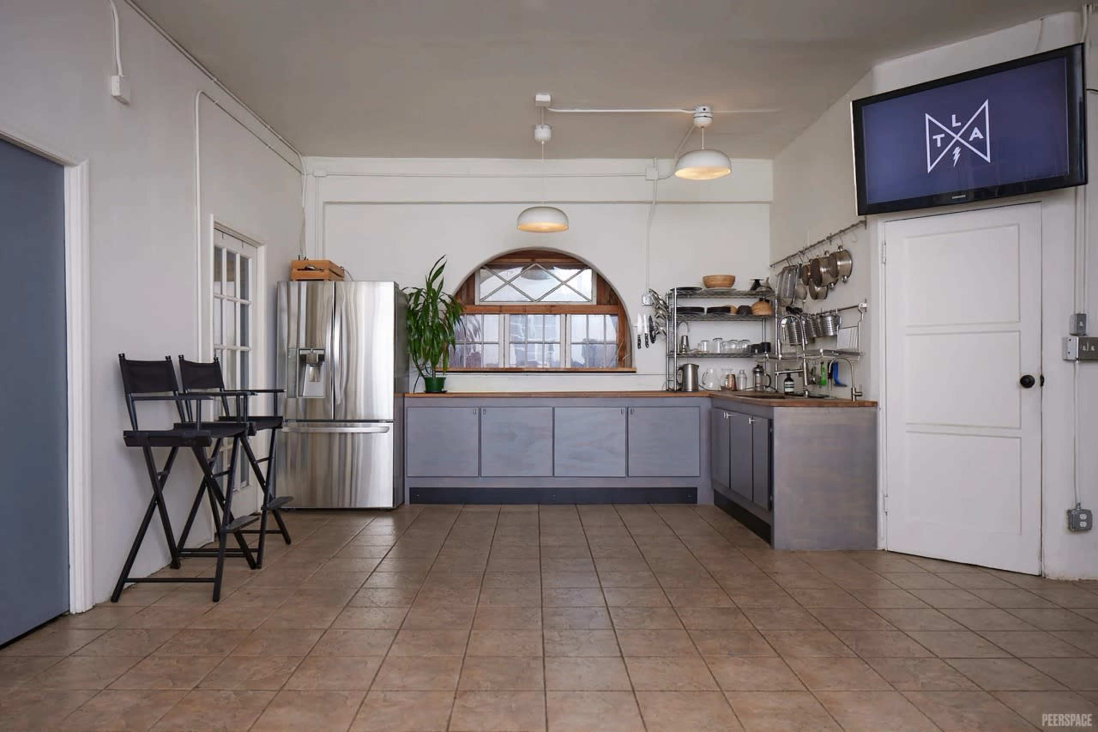 The image shows a modern kitchen space featuring a refrigerator, a minimalist countertop with utensils hanging, and two director's chairs positioned in front of a window.