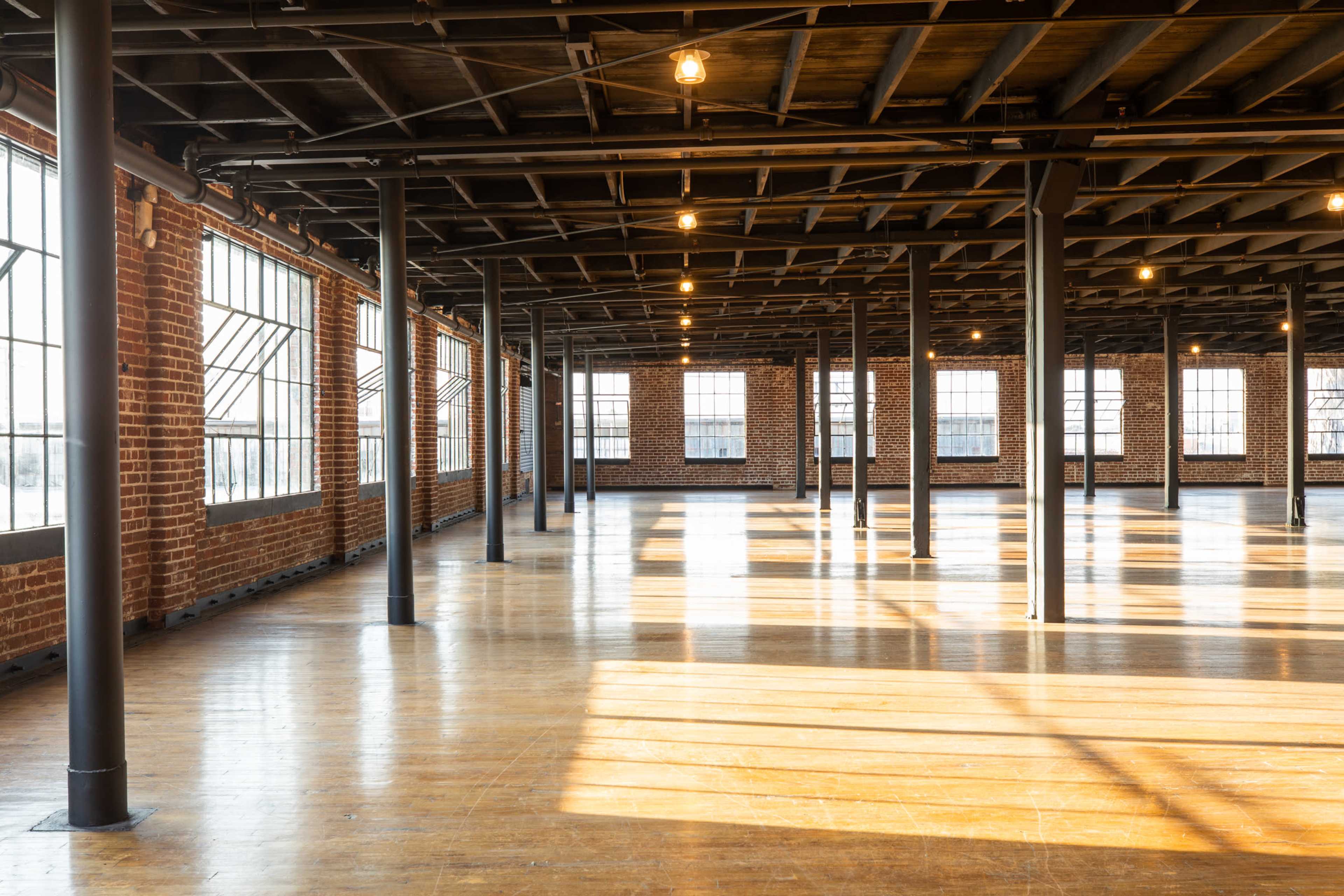 A spacious, industrial-style room with wooden floors and large windows, featuring exposed brick walls and metal support beams.