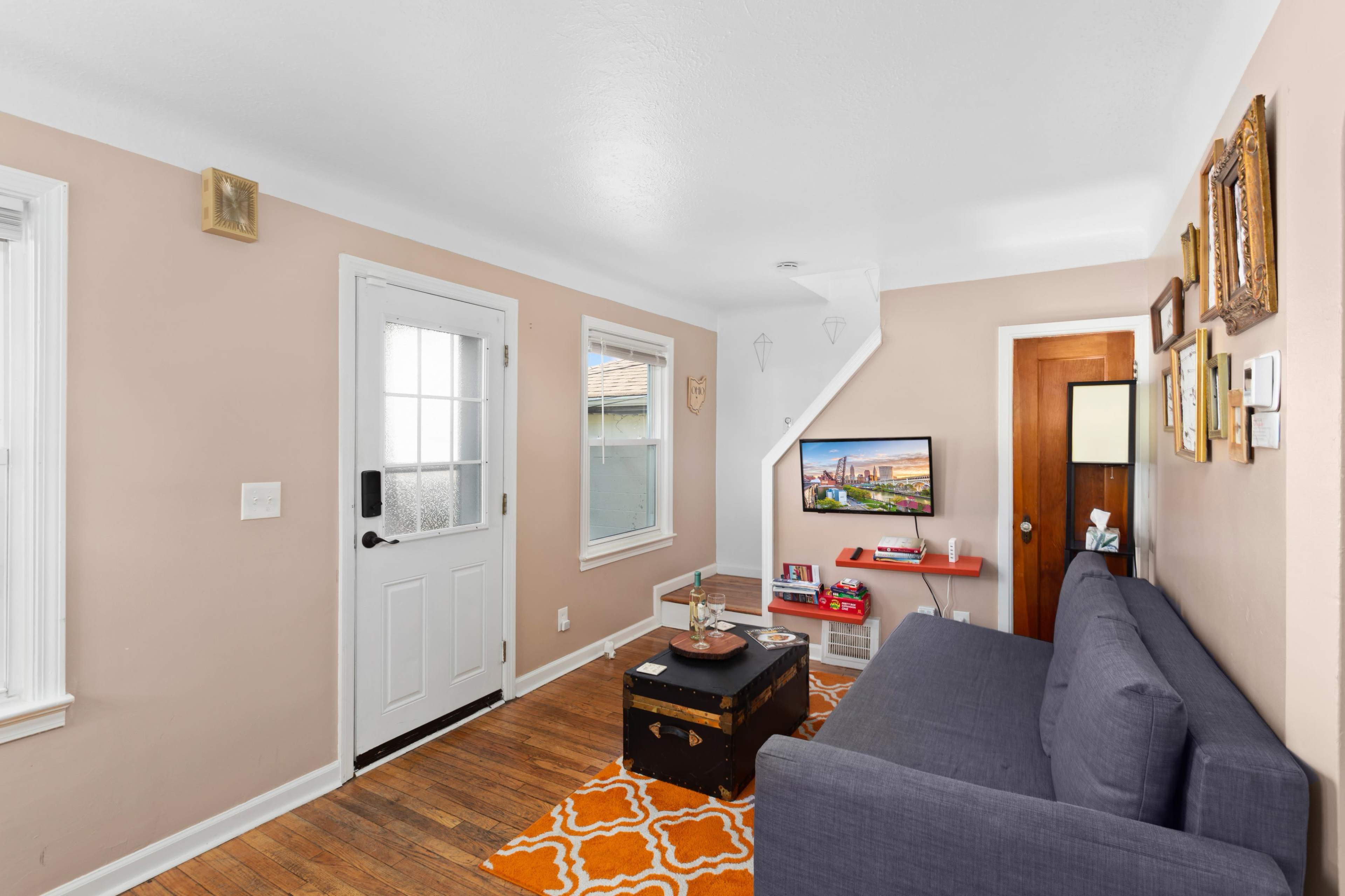 The living room features a gray sofa, an orange patterned rug, a small coffee table, and a television against the wall, with a front door and a staircase visible.
