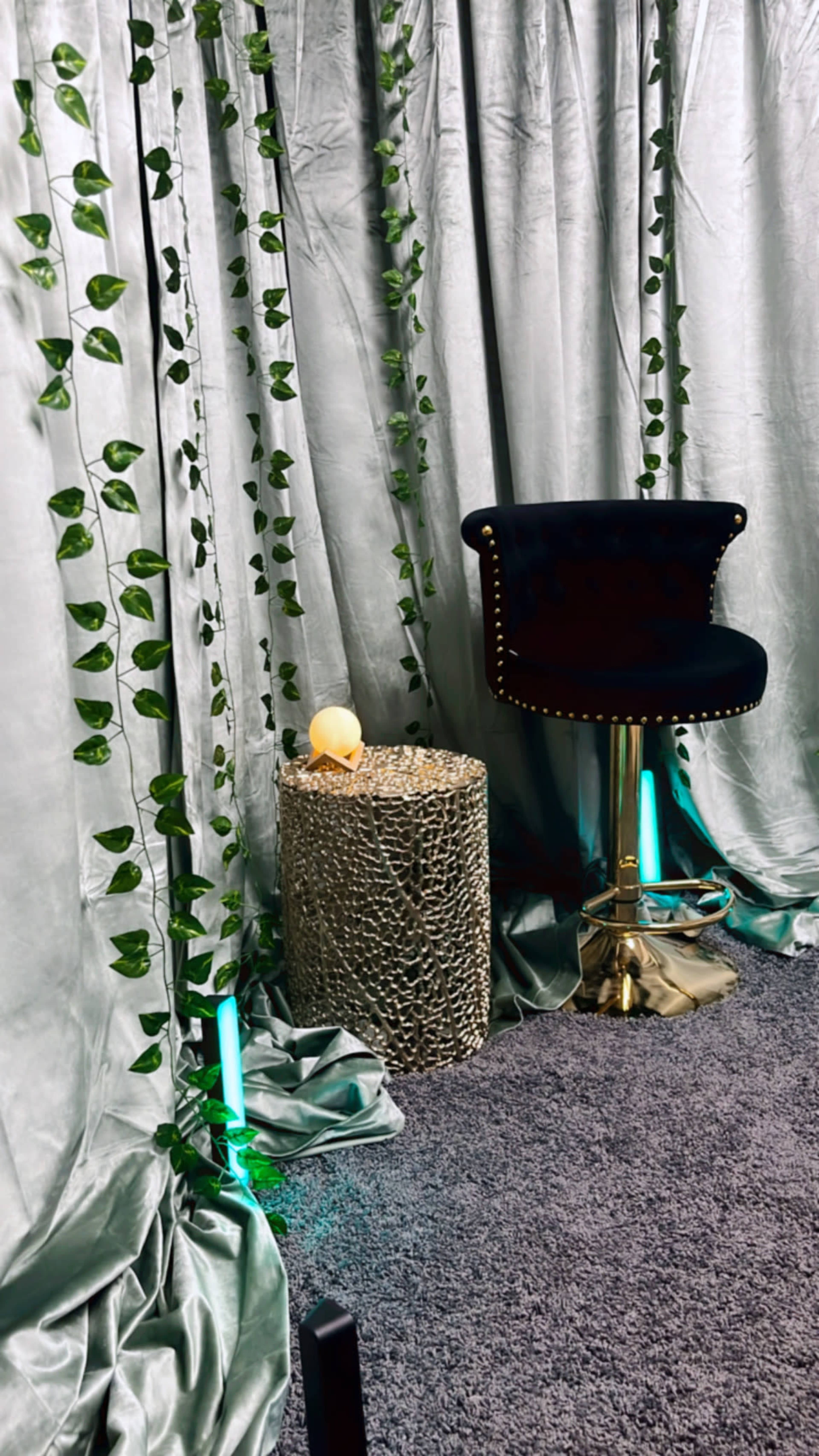 A black swivel chair next to a textured, metallic side table illuminated by teal lights, surrounded by silver curtains draped with green vines.