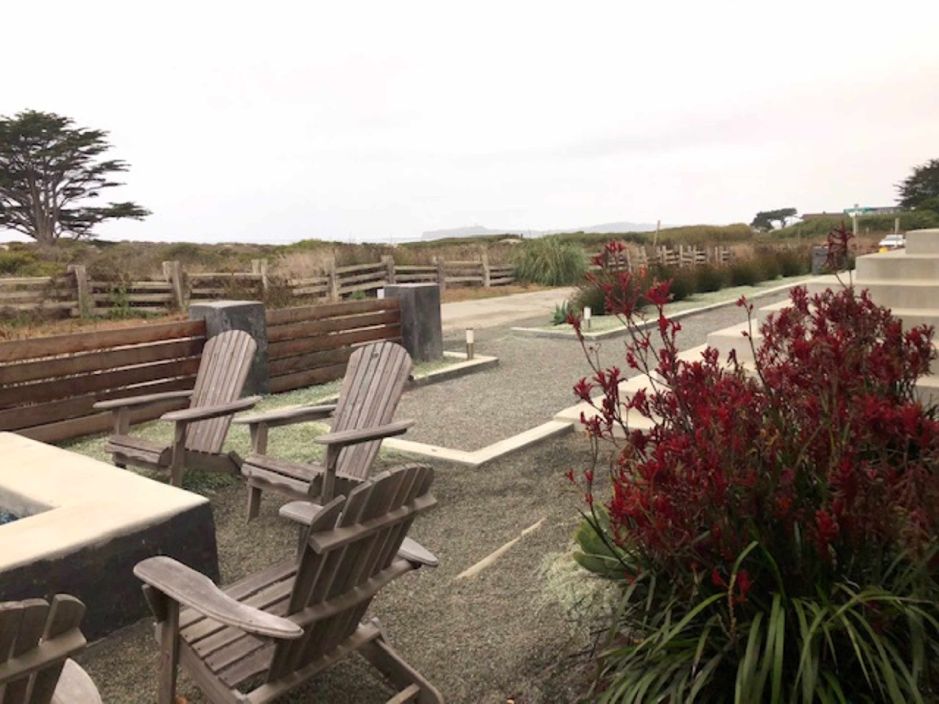 A landscaped outdoor area with wooden chairs, red plants, and pathways, set against a backdrop of grassy fields and a wooden fence.