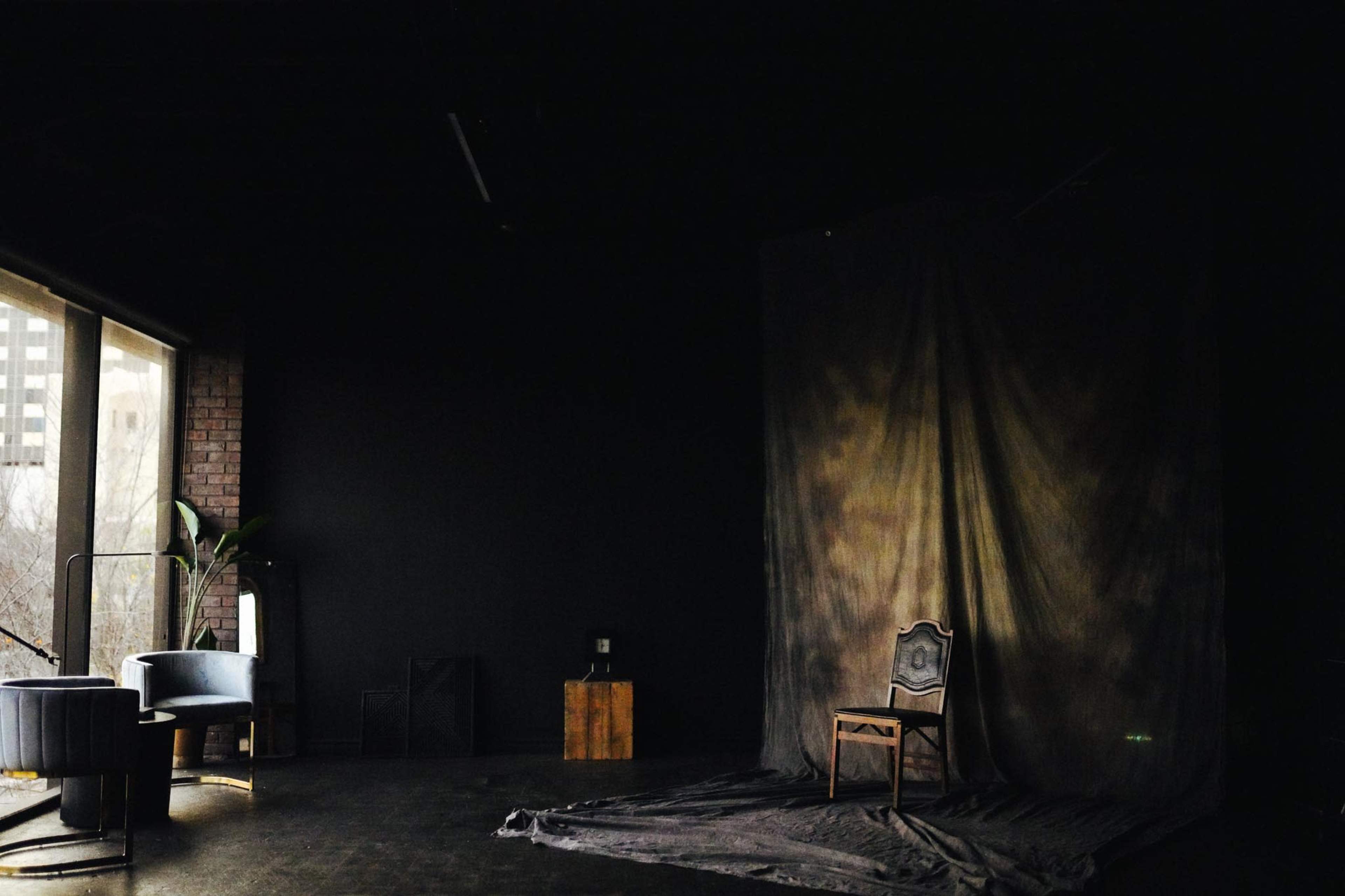 A dimly lit room features a chair in front of a draped backdrop with a window revealing a cityscape outside.