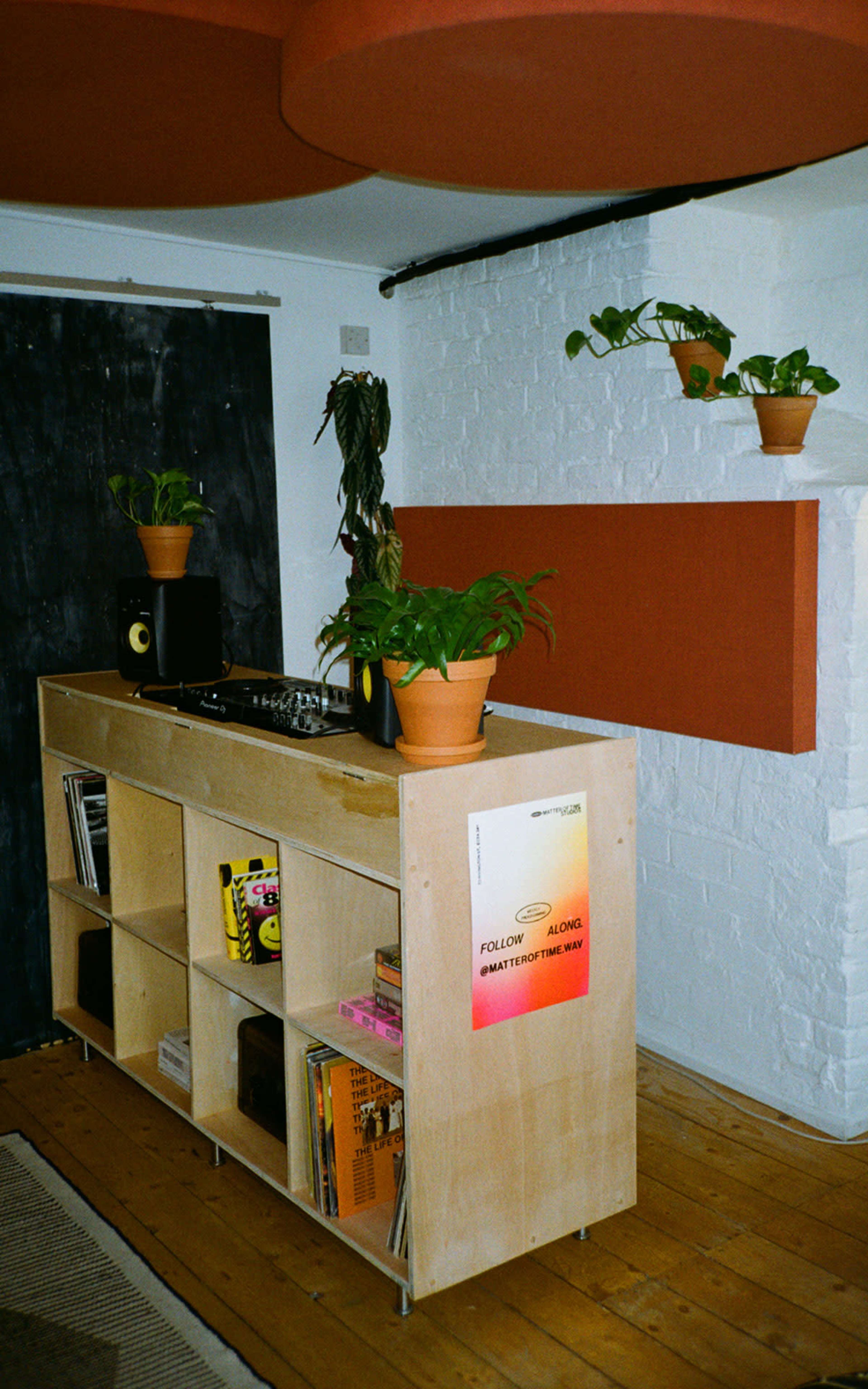 A wooden shelving unit holds vinyl records and a turntable, with potted plants placed on top in a minimalist room featuring a dark wall and circular orange ceiling elements.