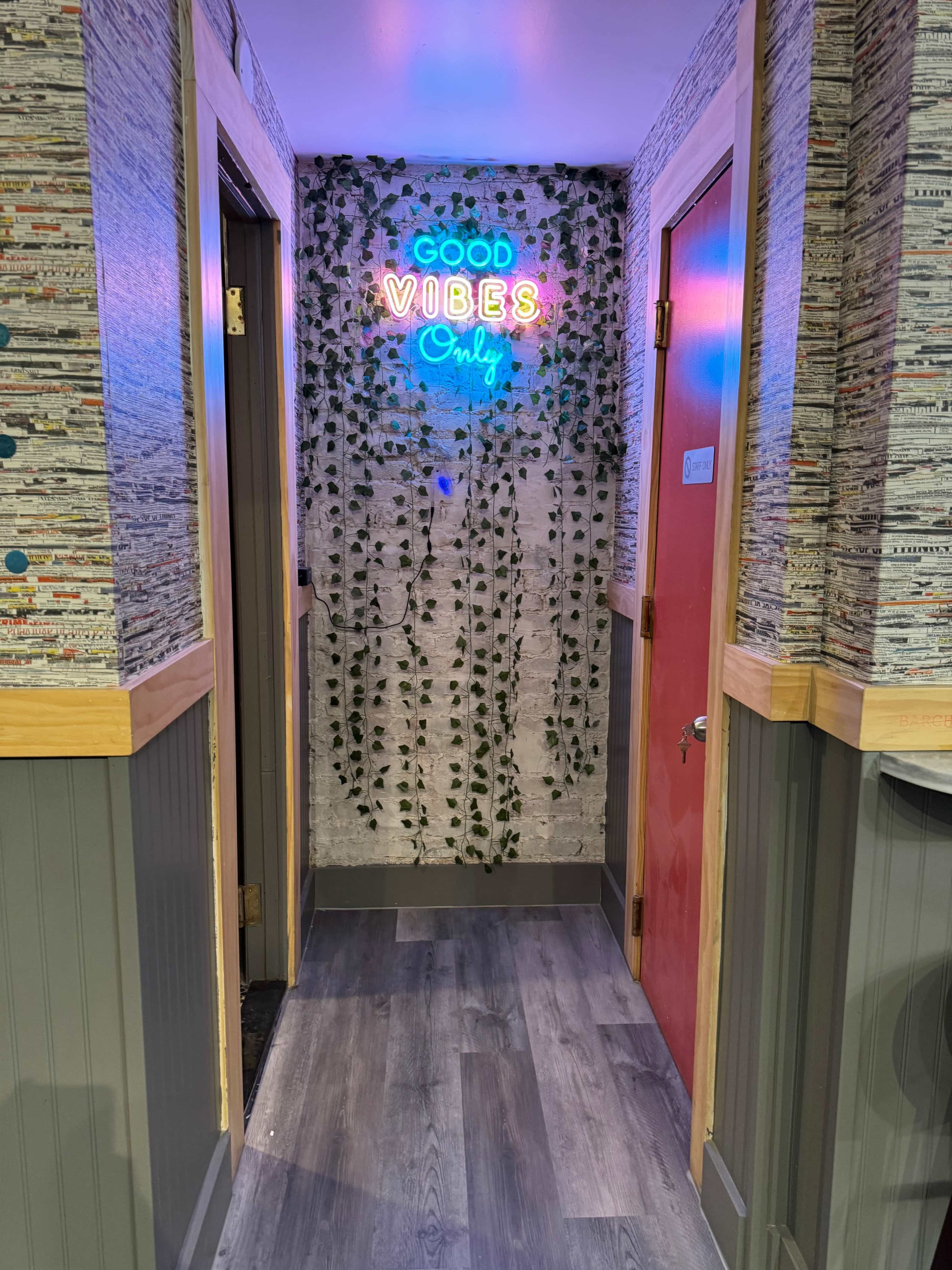 A narrow hallway with a neon sign reading "Good Vibes Only," surrounded by green vines on the wall and colorful wallpaper.