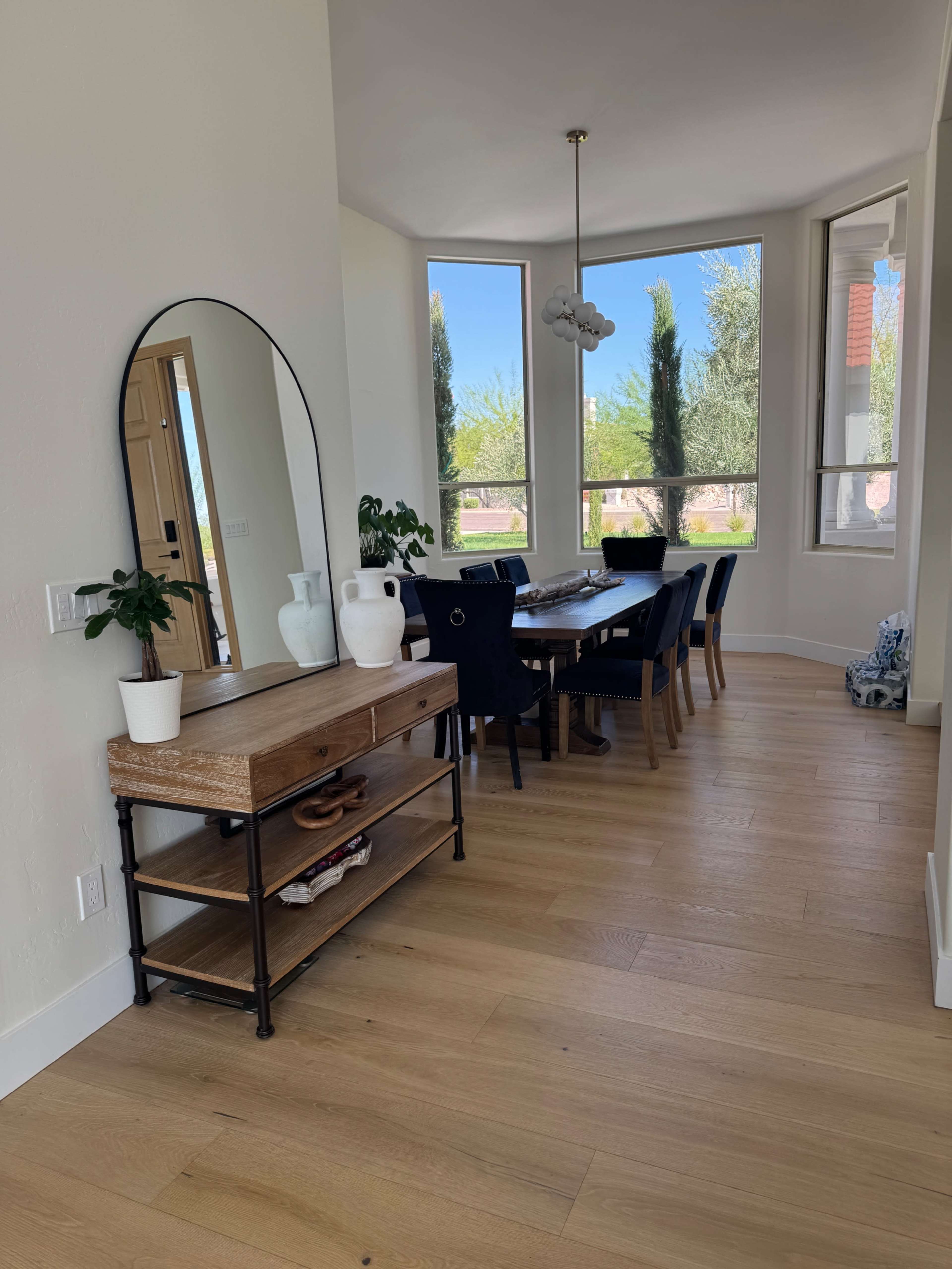 The image shows a dining area with a wooden table and black chairs, a console table with a mirror and plants, and large windows allowing natural light.