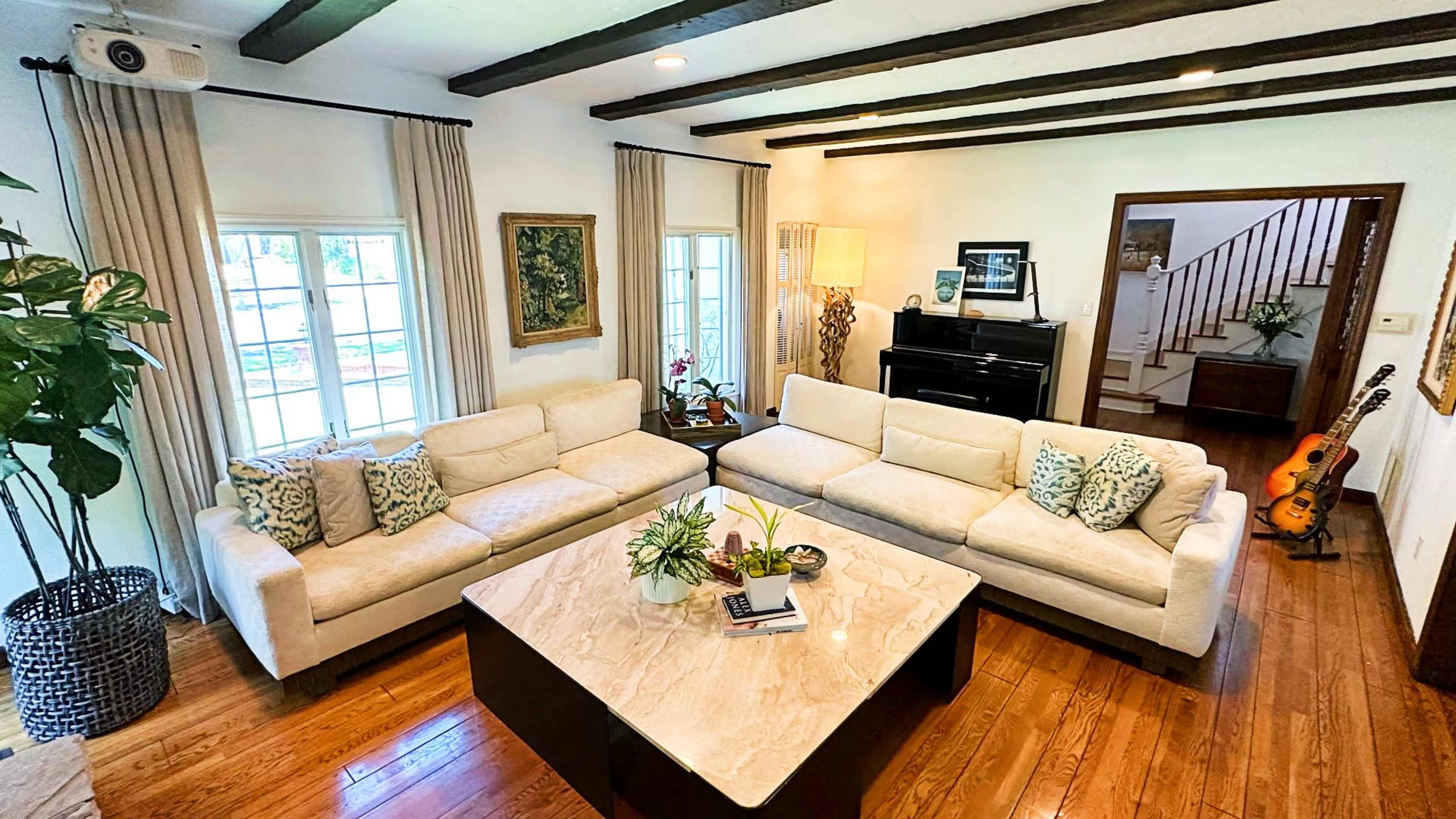 A living room with two white sofas arranged around a coffee table, a piano in the corner, and a staircase leading to another floor.