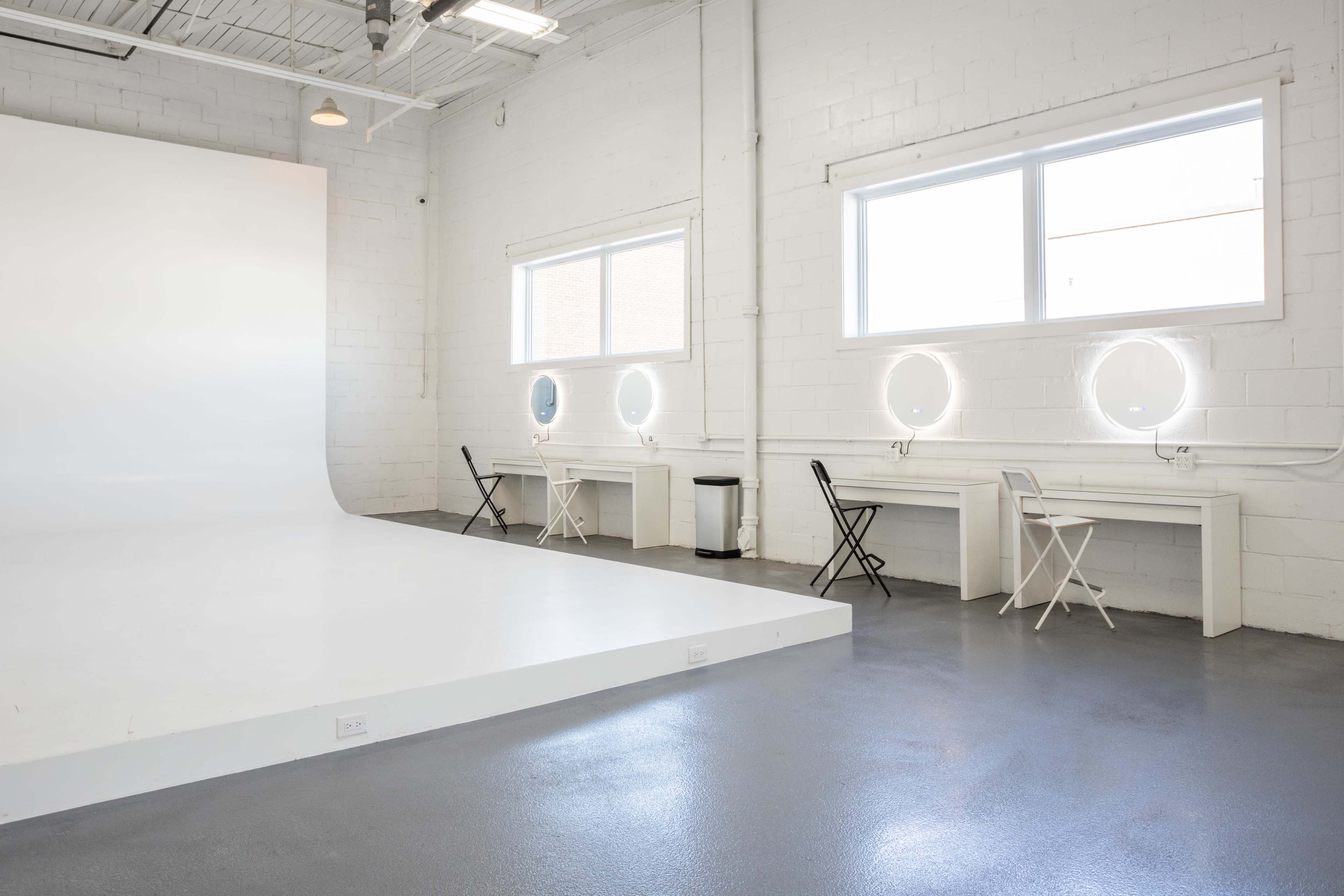 The image shows a photography studio with a white backdrop, two circular mirrors with lights, and two tables with chairs positioned against the walls.