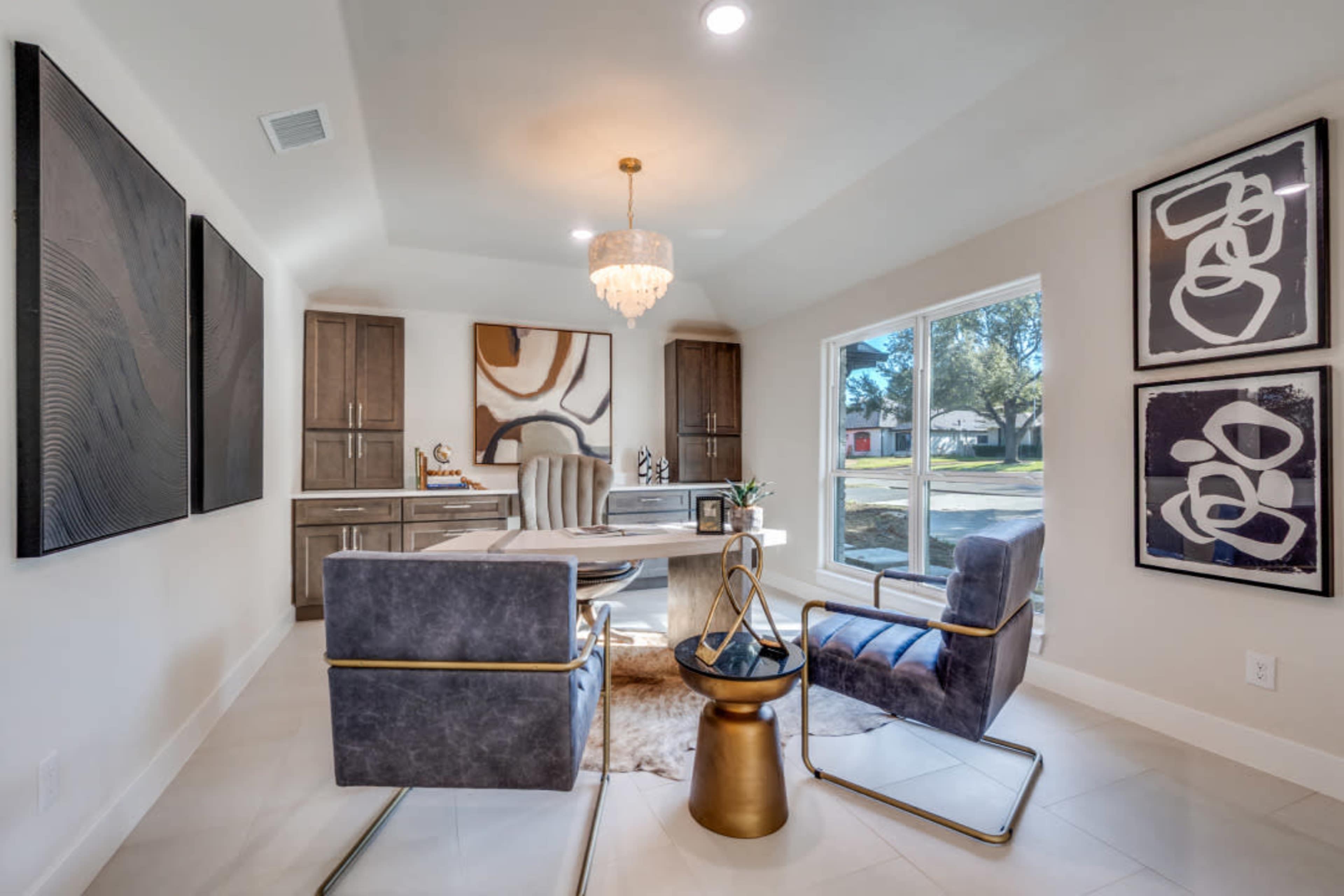 A modern office space features a sleek table and stylish chairs, illuminated by a chandelier, with abstract artwork on the walls and a view of the outdoors.