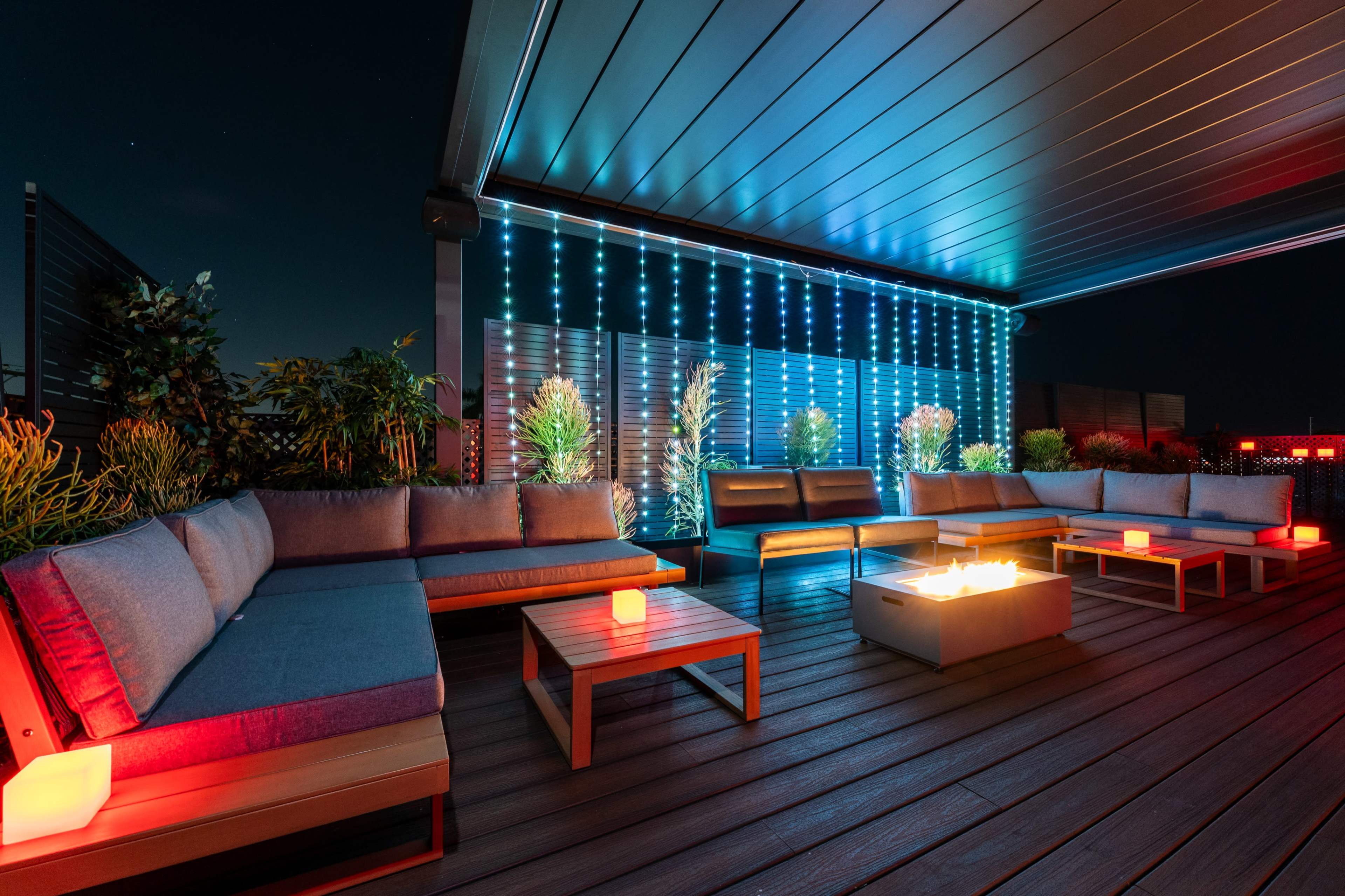 The image shows a rooftop lounge area featuring modern seating, illuminated plants, and a fire pit, all under a night sky.