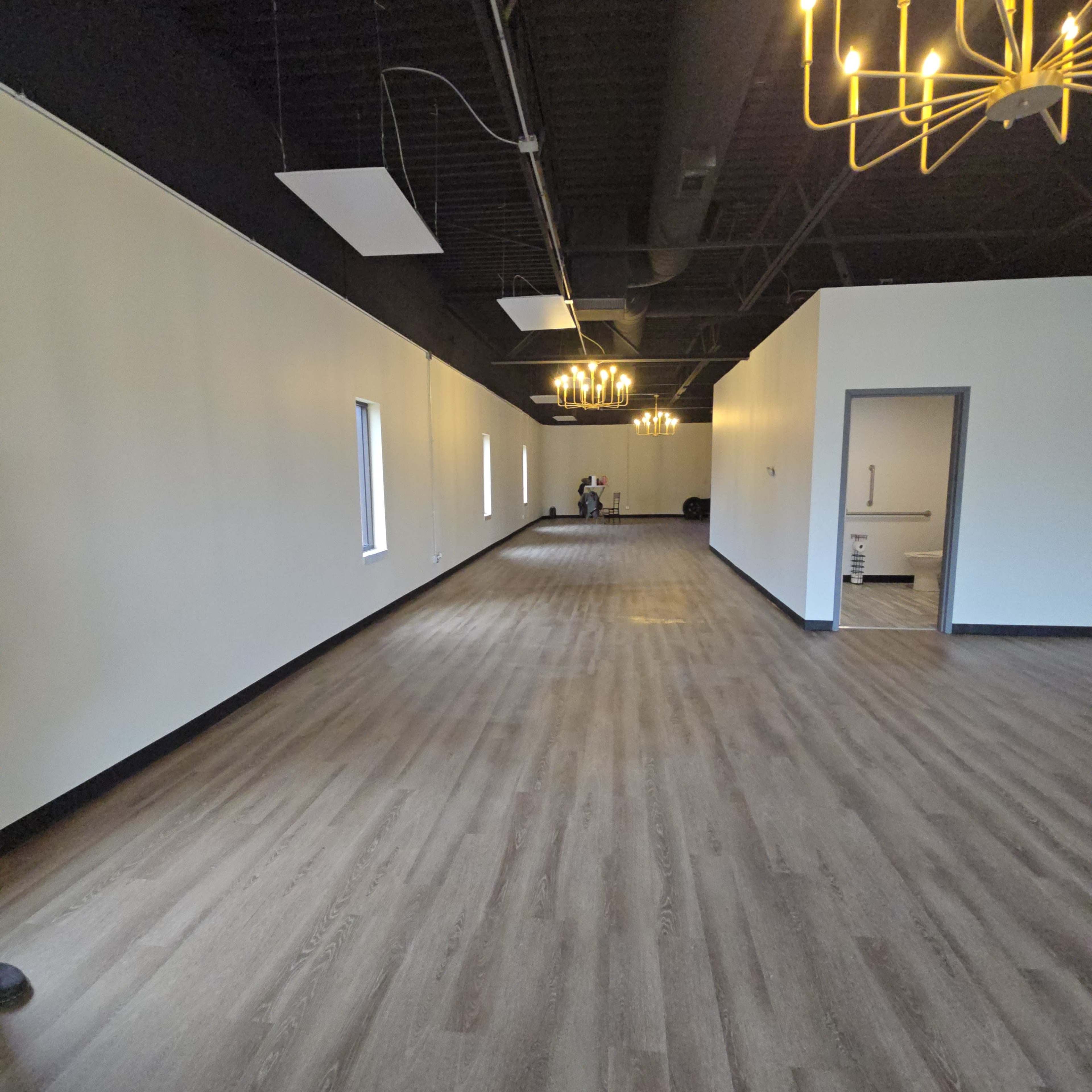 The image shows a spacious, newly constructed interior with wooden flooring, white walls, and modern light fixtures hanging from the ceiling.