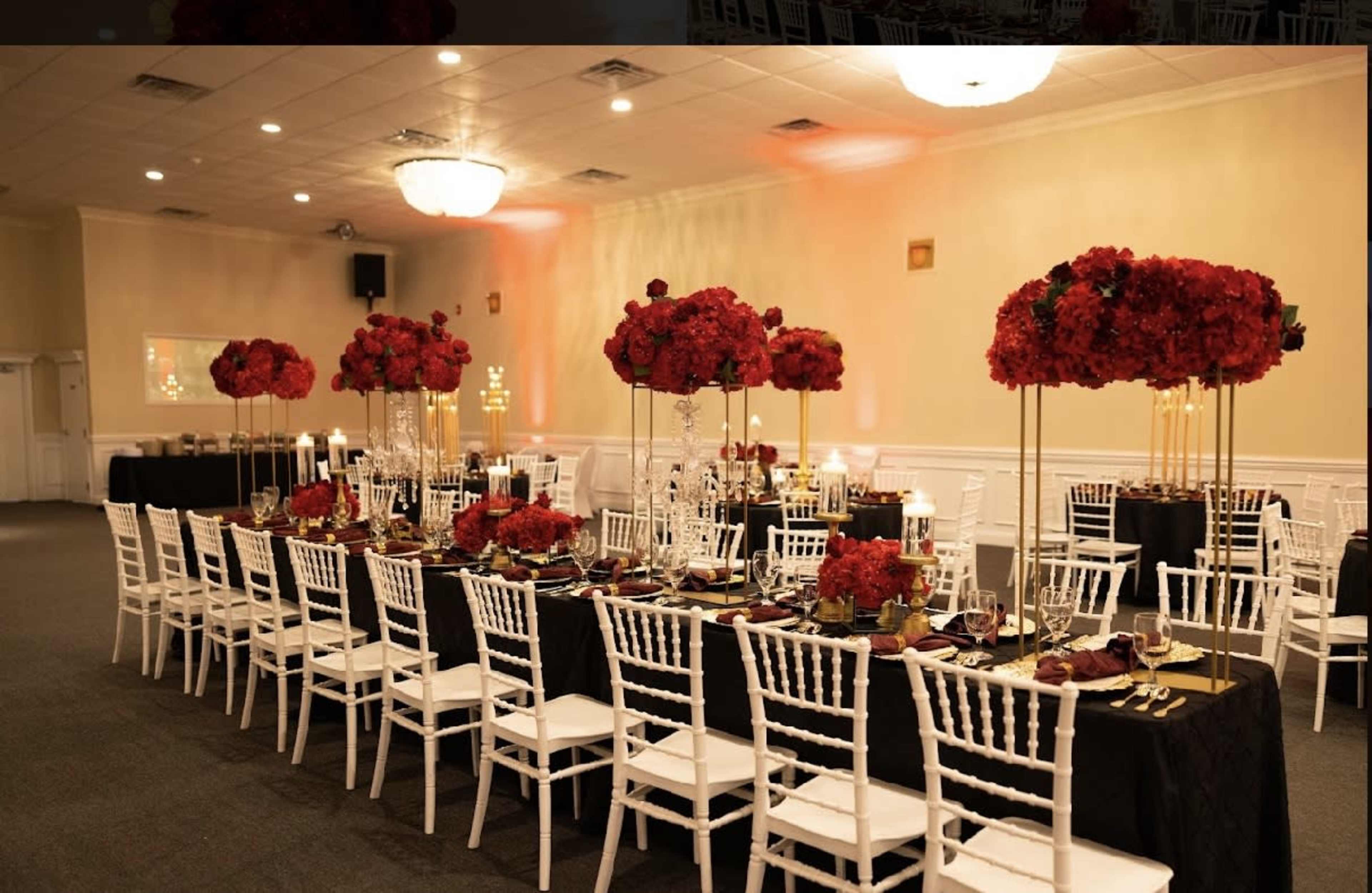 A elegantly arranged dining area with long tables set for an event, featuring white chairs, black tablecloths, and tall centerpieces of red flowers.