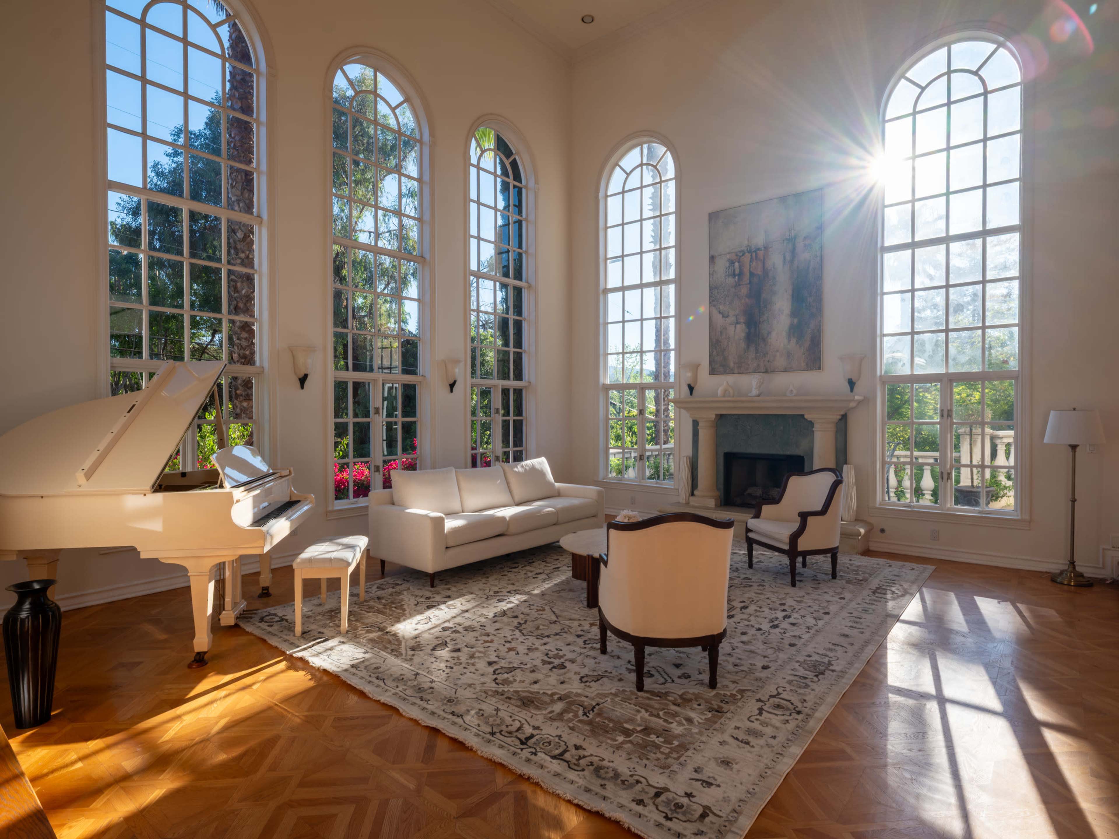 A bright, spacious living room features large arched windows, a grand piano, a white sofa, and an elegant armchair, all accentuated by natural light streaming in.