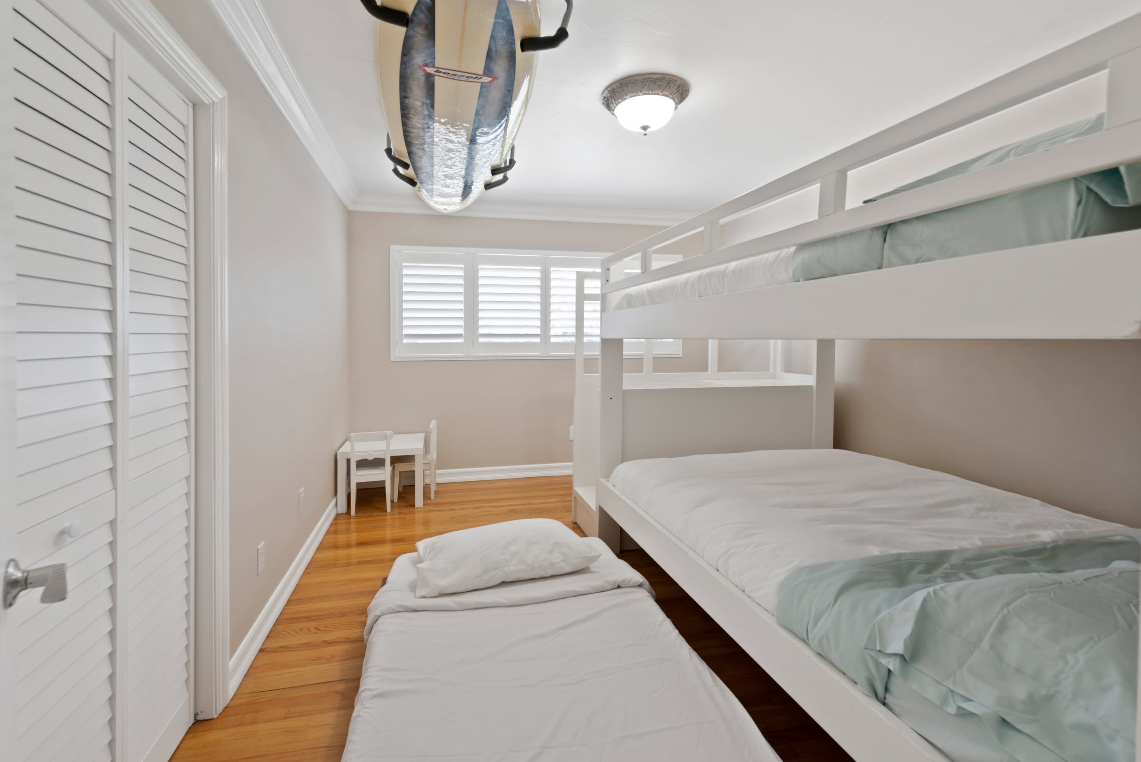 The room features a bunk bed, a mattress on the floor, and a surfboard mounted on the ceiling, with light-colored walls and hardwood floors.