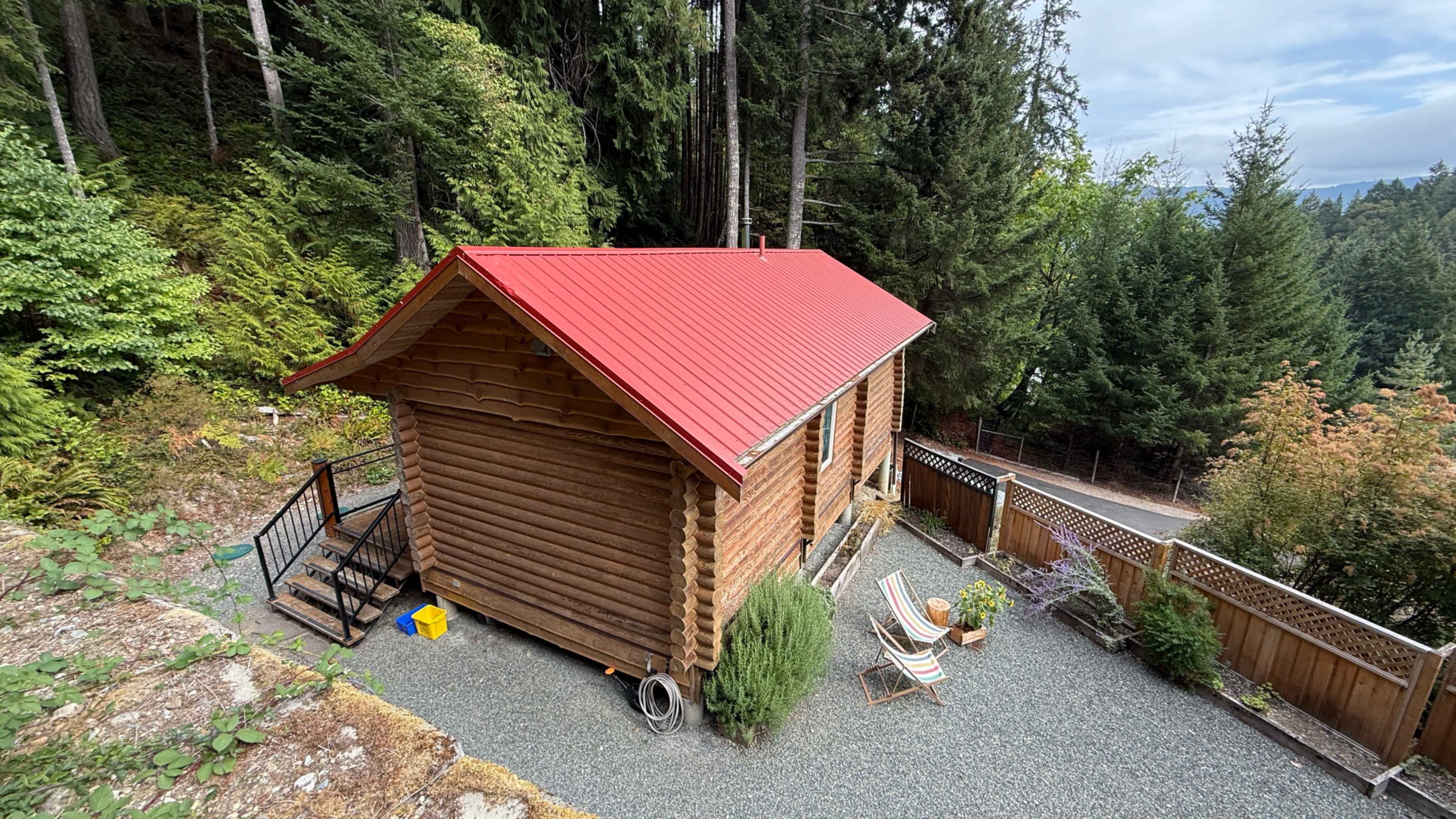 A wooden cabin with a red metal roof sits surrounded by trees and gravel, featuring a small staircase and outdoor seating.