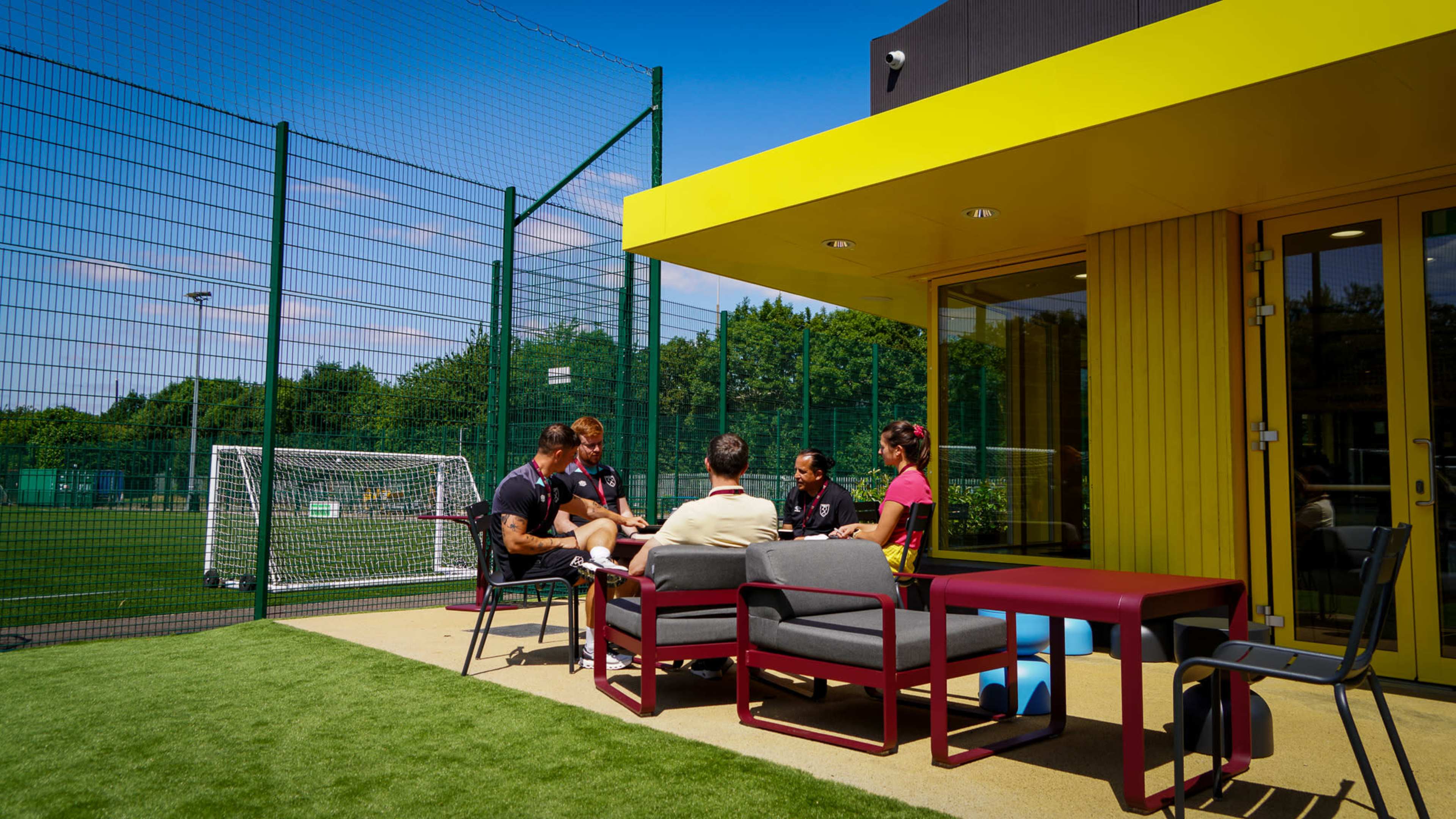 A group of five people is seated outside a brightly colored building near a soccer field, discussing while surrounded by a fence and goalpost.