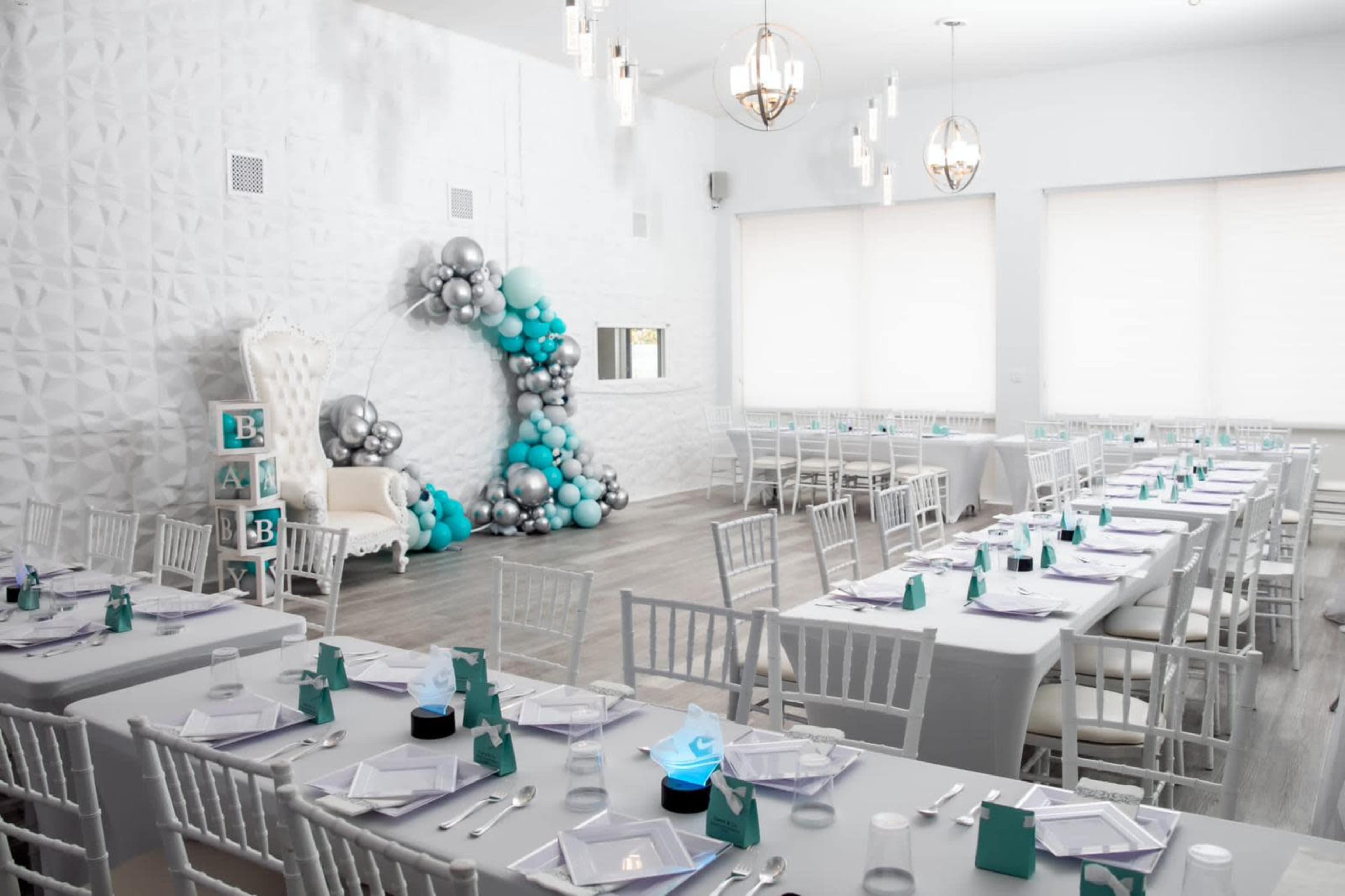 A bright event space arranged with white tables and chairs, featuring a decorative balloon arch and a throne-like chair at one end.
