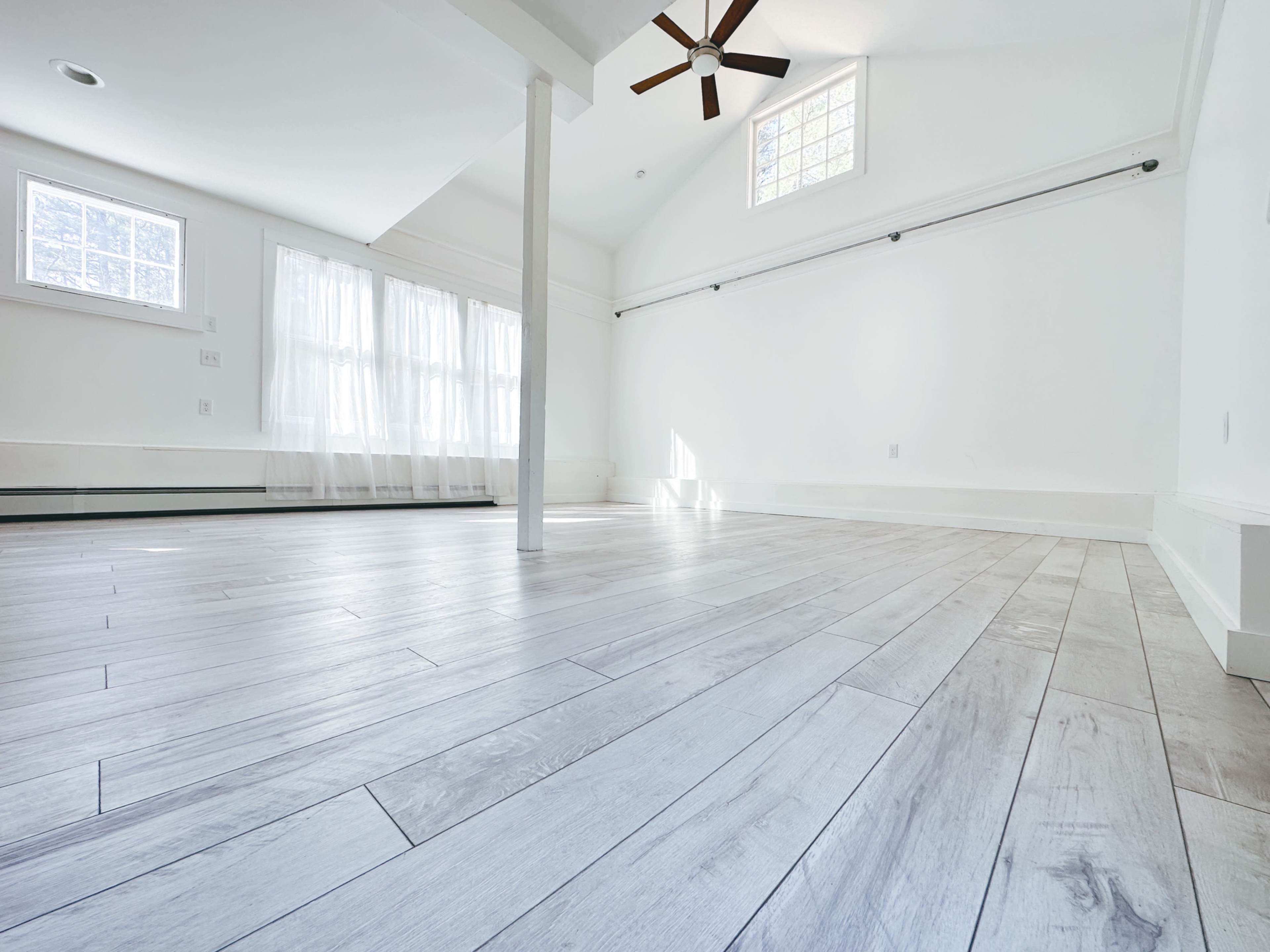 A bright, empty room with large windows, white walls, and light-colored wooden flooring.