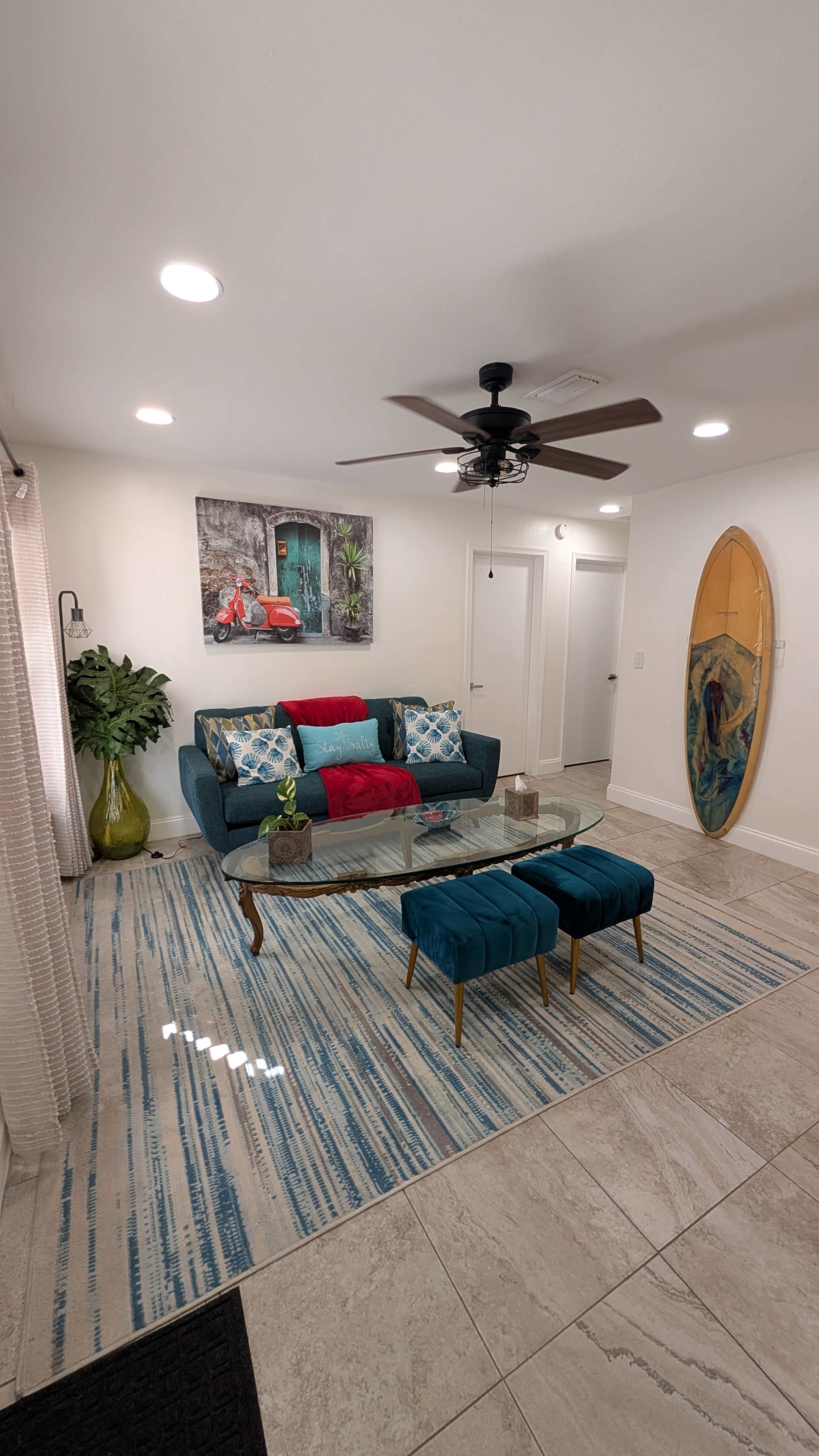 The scene features a living room with a blue couch, colorful throw pillows, a glass coffee table, and two teal ottomans on a striped area rug, alongside a surfboard mounted on the wall.