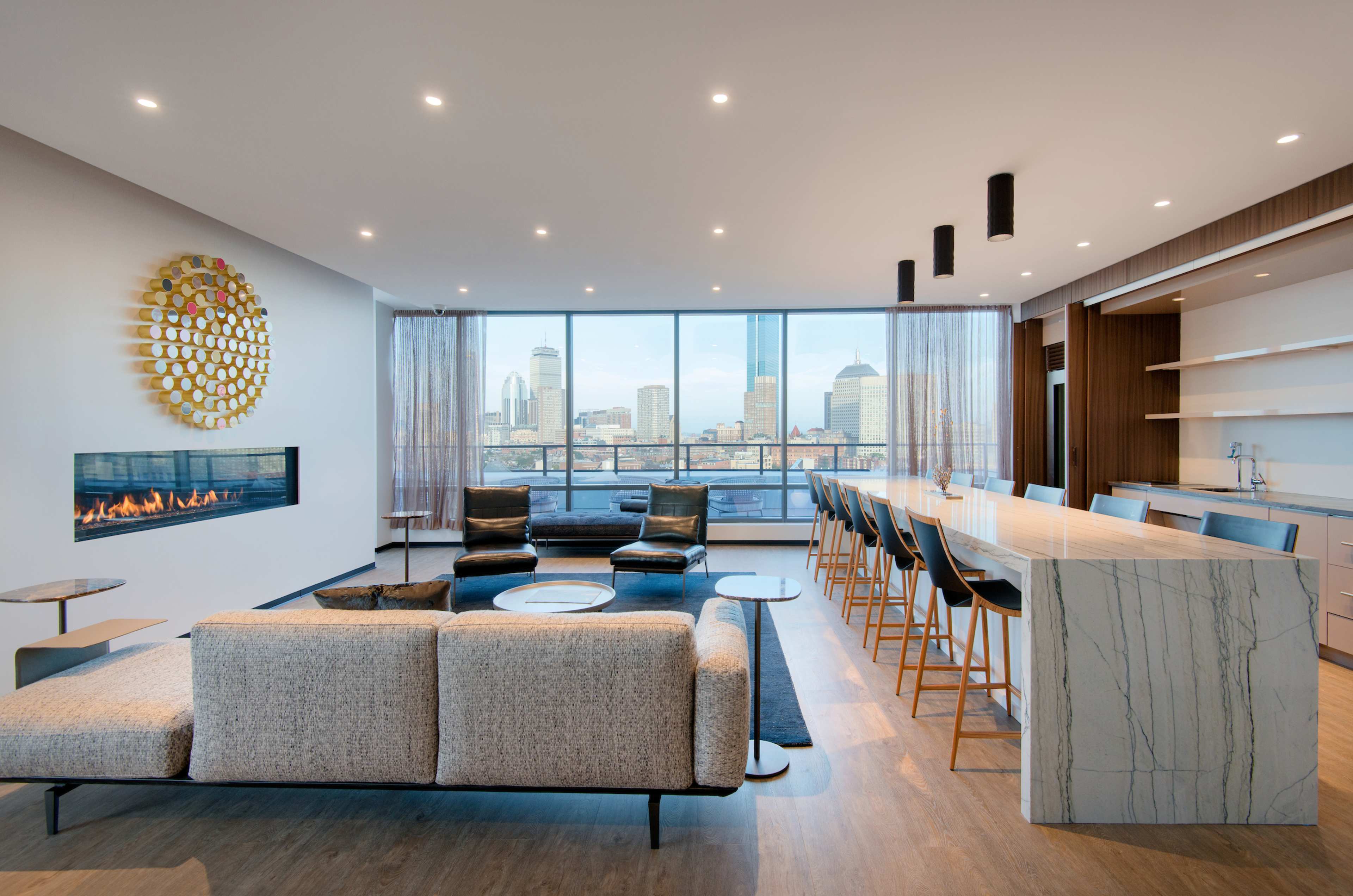 A modern living room features a sectional sofa and a bar-style seating area, with large windows offering a cityscape view and a linear fireplace along one wall.