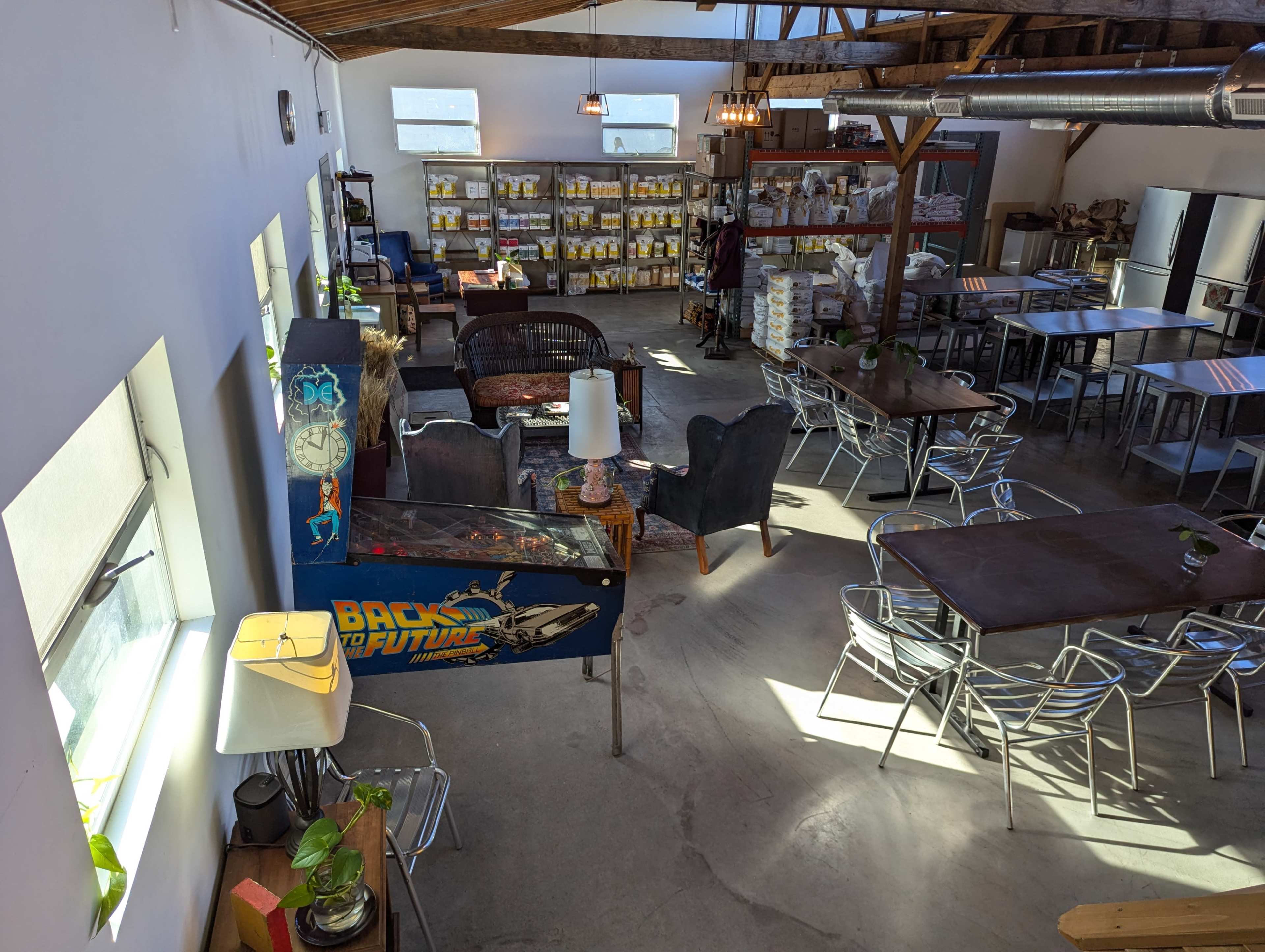 A spacious cafe with metal tables, chairs, a seating area, and shelves filled with packaged goods in the background.