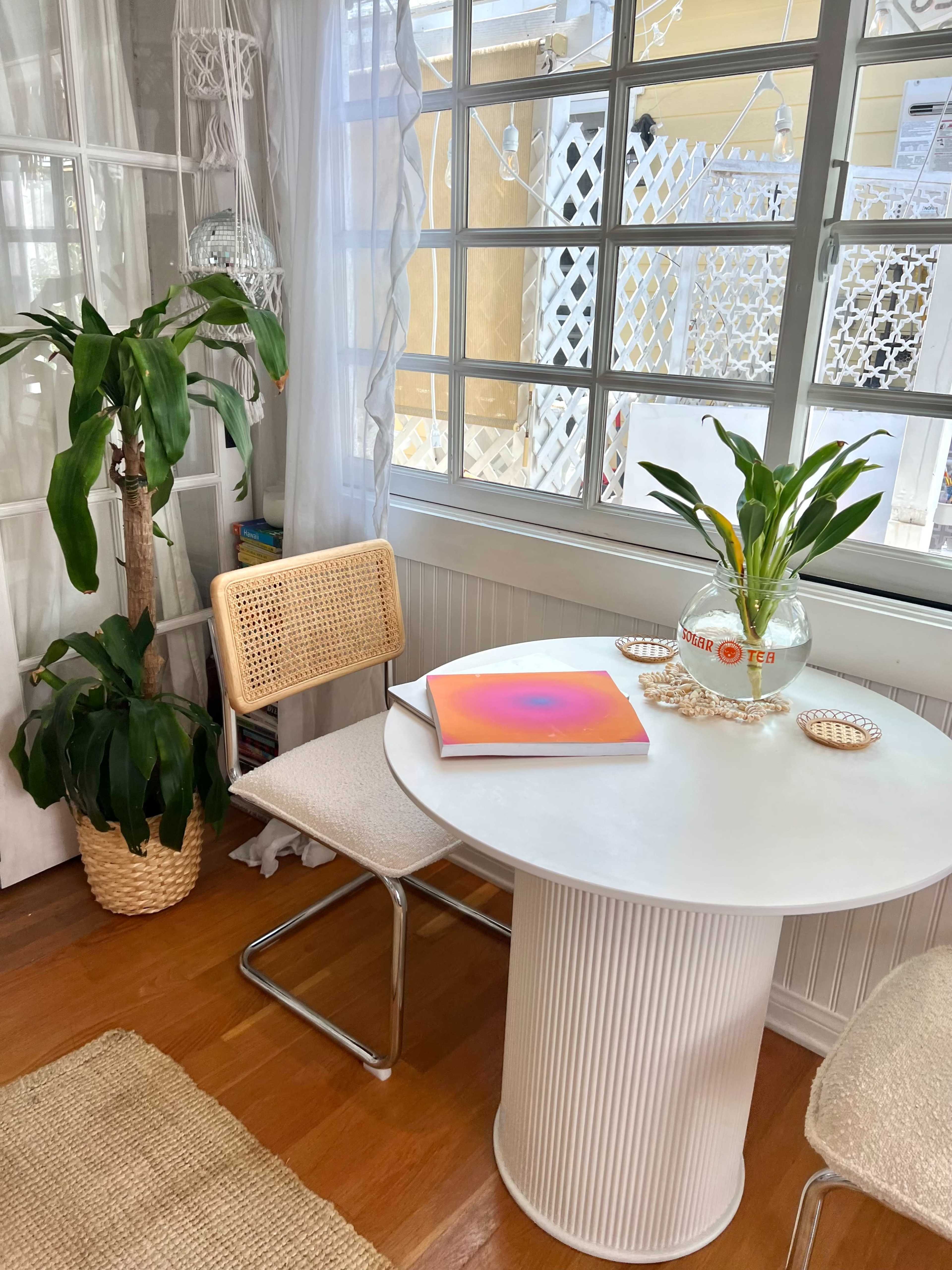 A round white dining table with a colorful book on top is set next to a chair and a potted plant near a window.