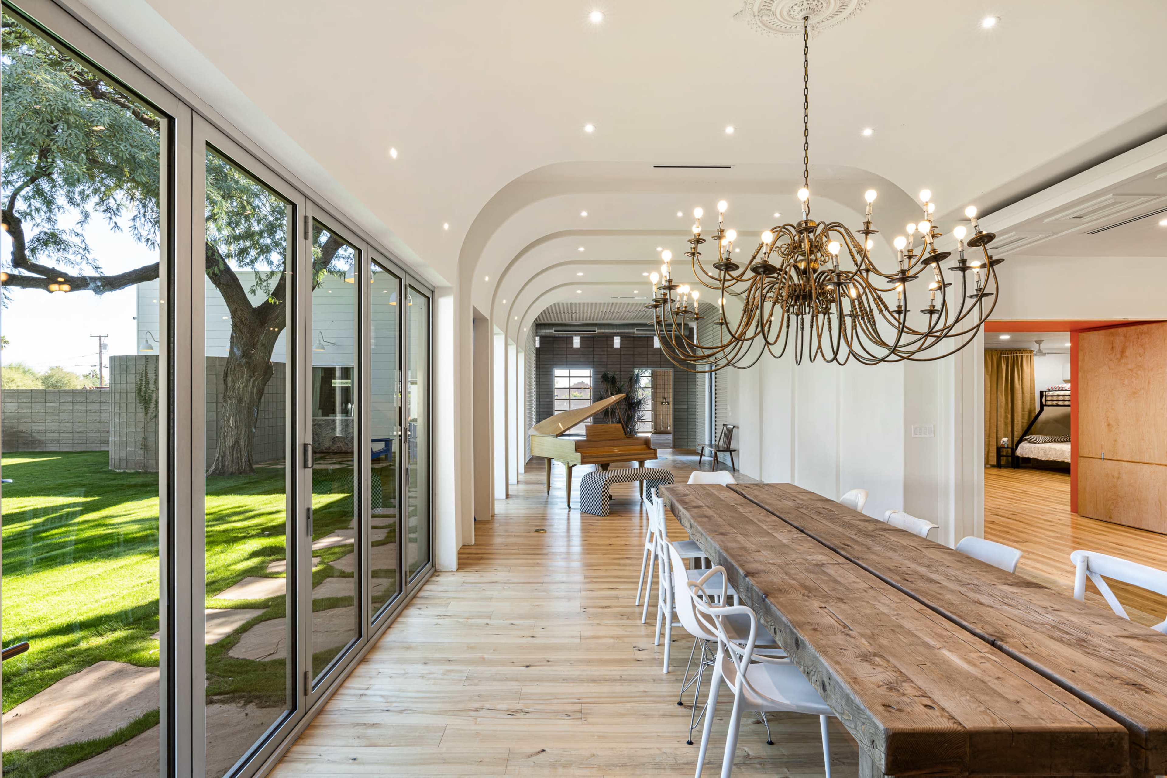 A dining area with a long wooden table, surrounded by white chairs, large glass windows overlooking a green lawn, and a grand piano in the corner.