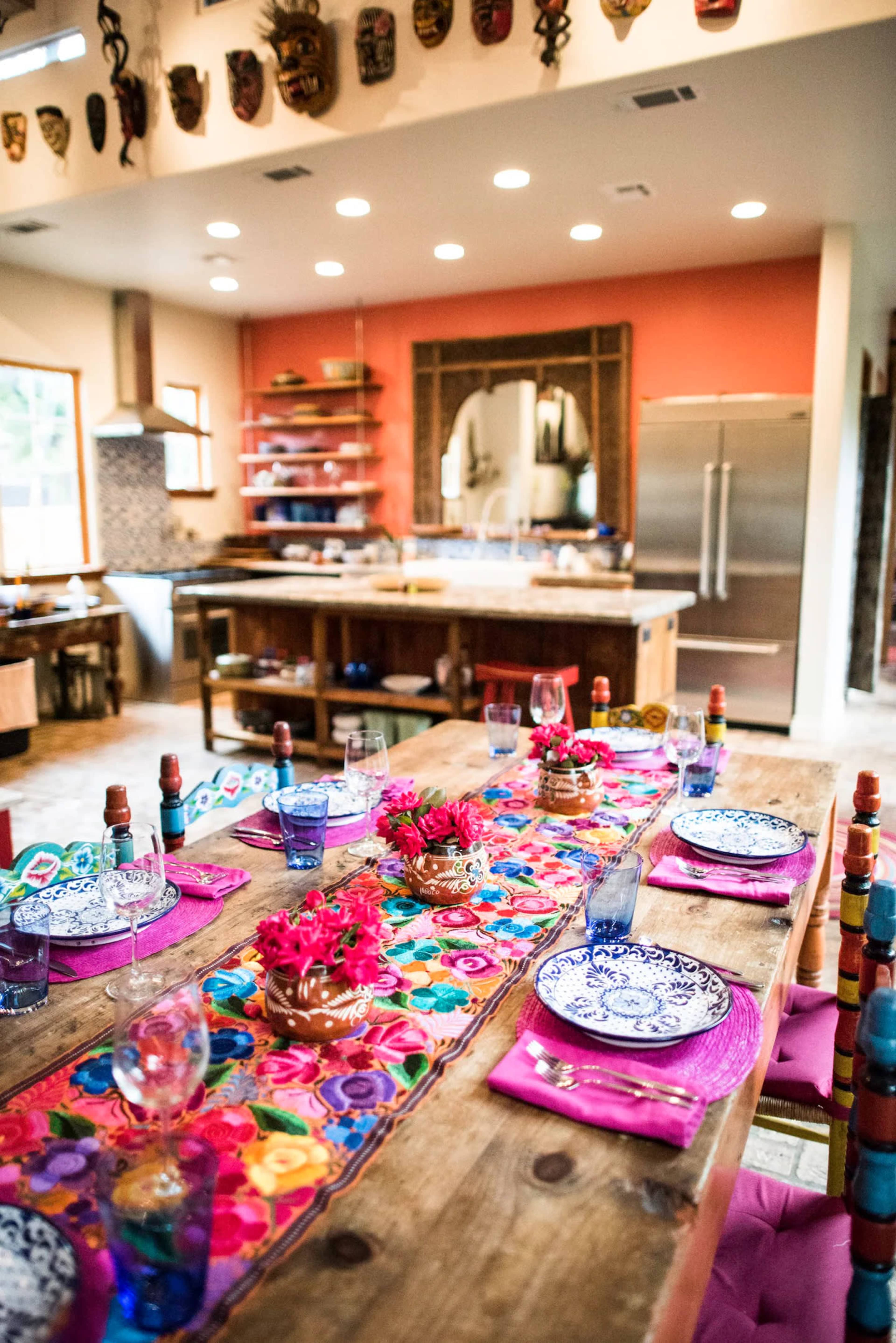 A brightly decorated dining table with colorful plates and glasses is set in a spacious kitchen featuring wooden shelves and modern appliances.