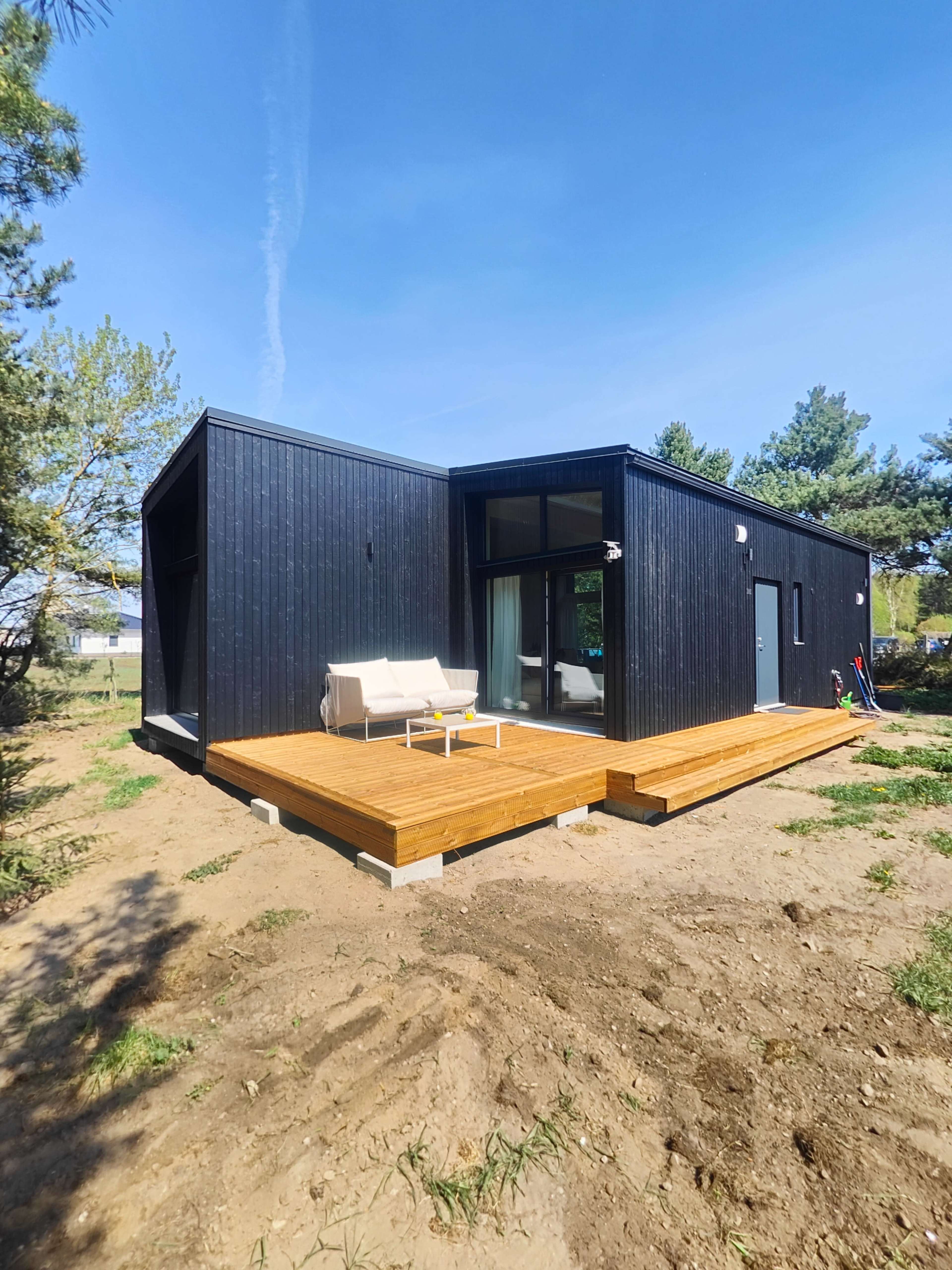 A modern black house with a wooden deck is set in a grassy area surrounded by trees under a clear blue sky.