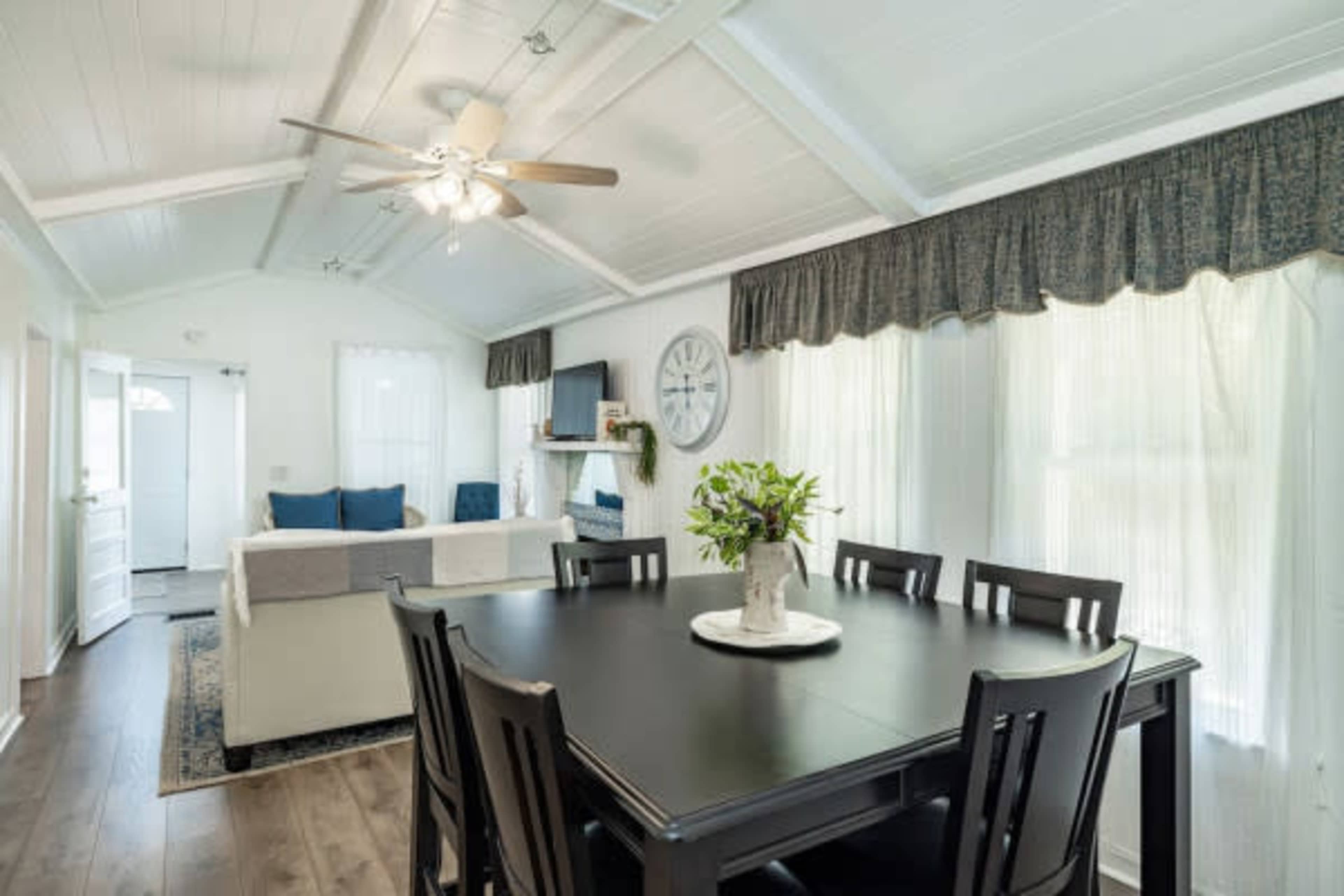 The image depicts a bright, open living area featuring a dining table in the foreground, a sofa and television in the background, and large windows with sheer curtains.