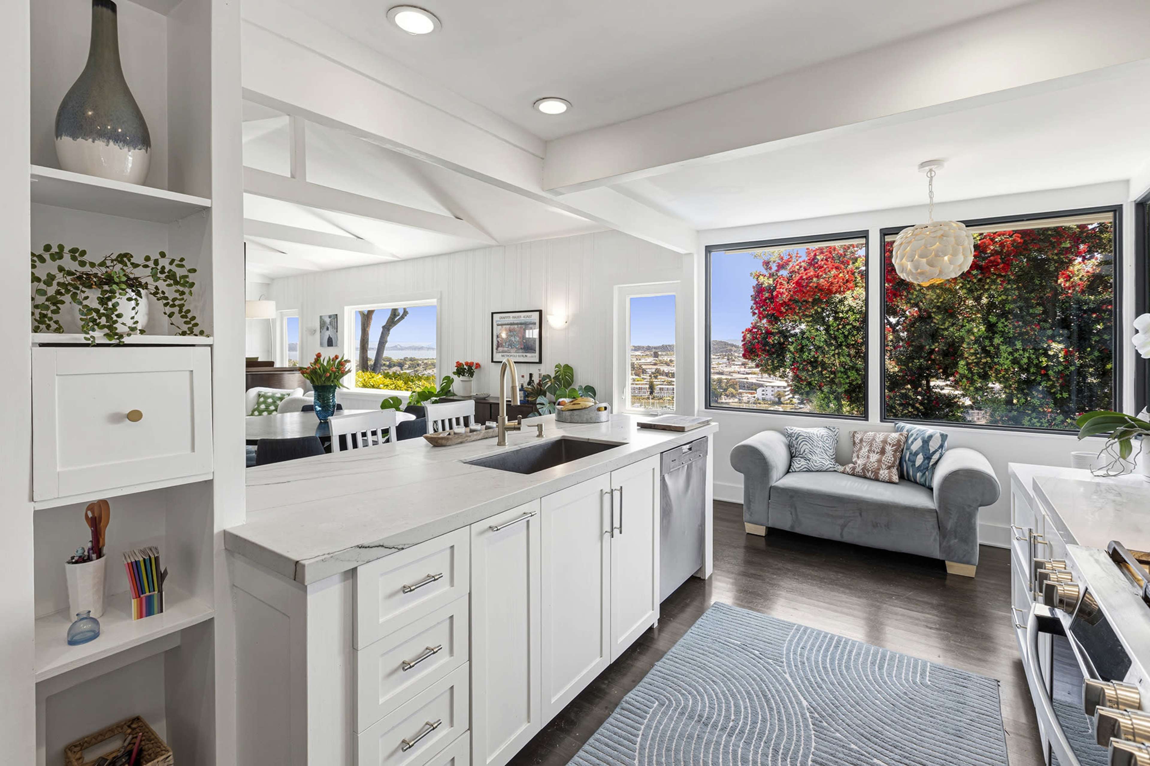 A modern kitchen features white cabinetry, a gray countertop, and large windows overlooking a vibrant garden.