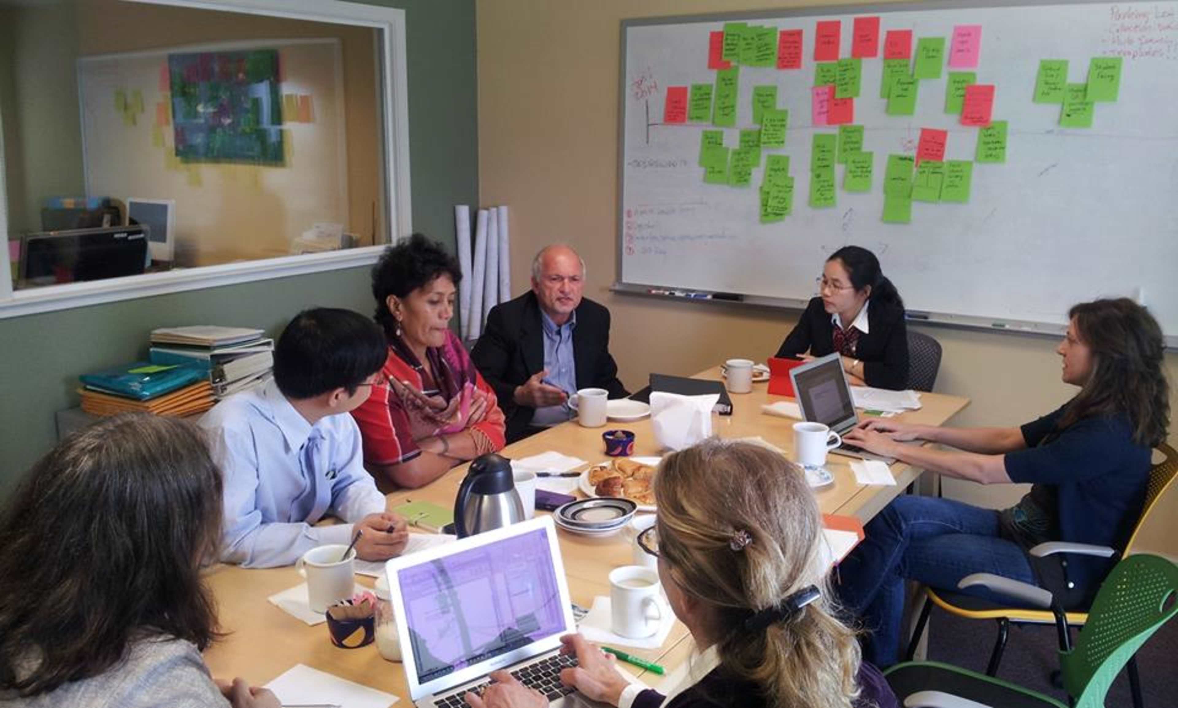 A group of eight people are seated around a conference table, engaged in discussion, with a whiteboard covered in colorful sticky notes visible in the background.