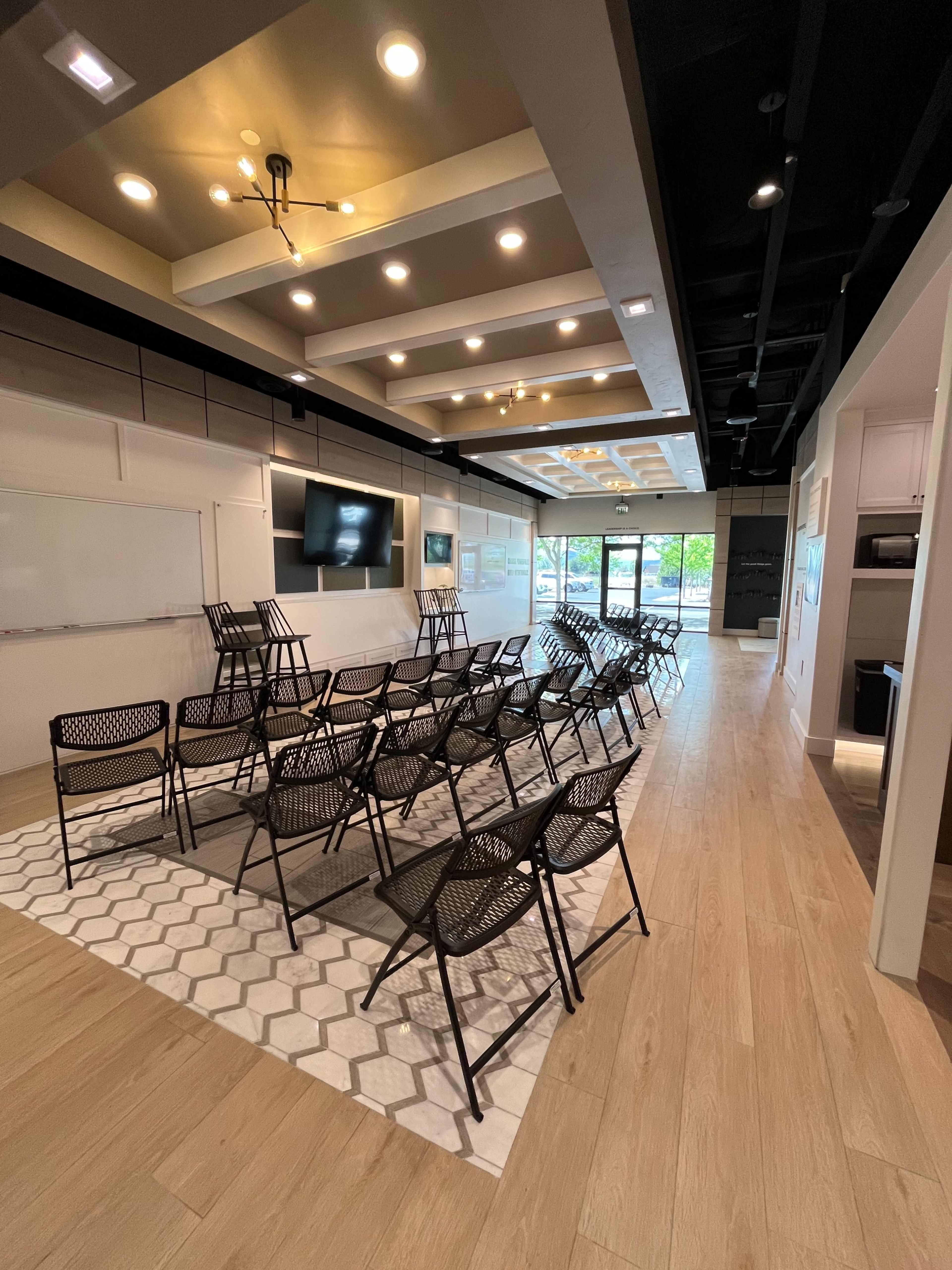The image shows a modern conference room arranged with black chairs facing a presentation area, featuring a large window allowing natural light to enter.