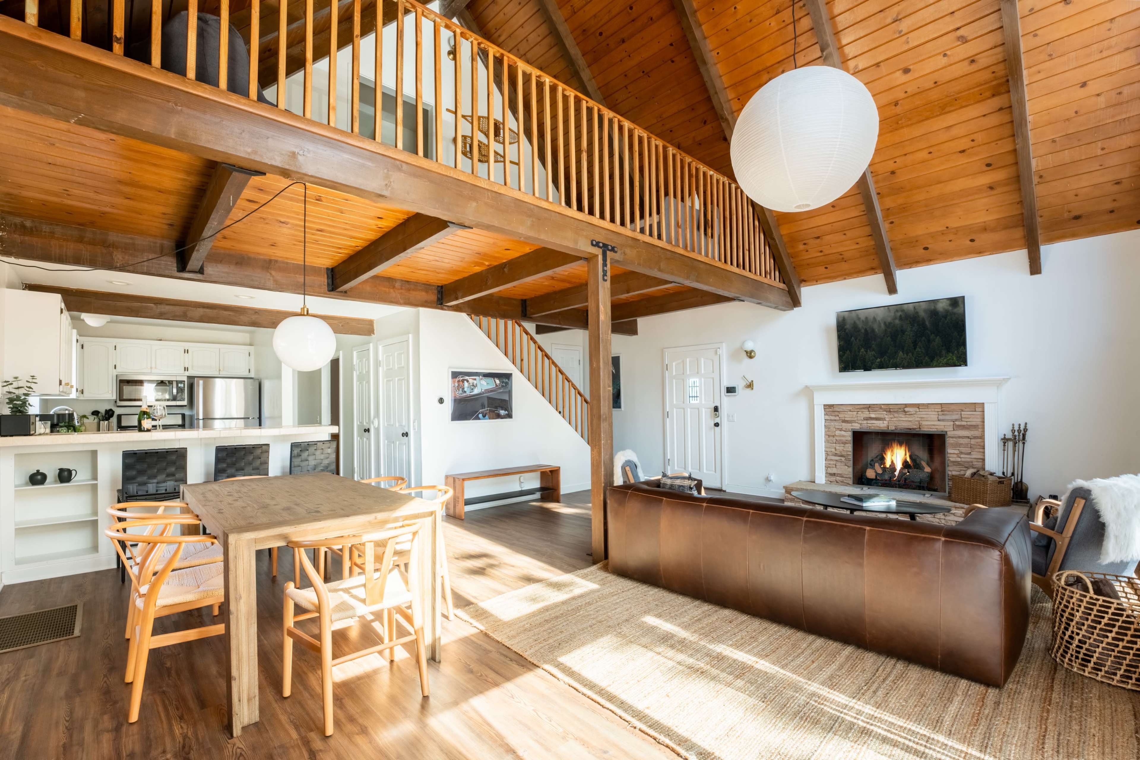 The image shows a spacious living area with high wooden ceilings, a fireplace, and a dining table near a staircase leading to an upper level.